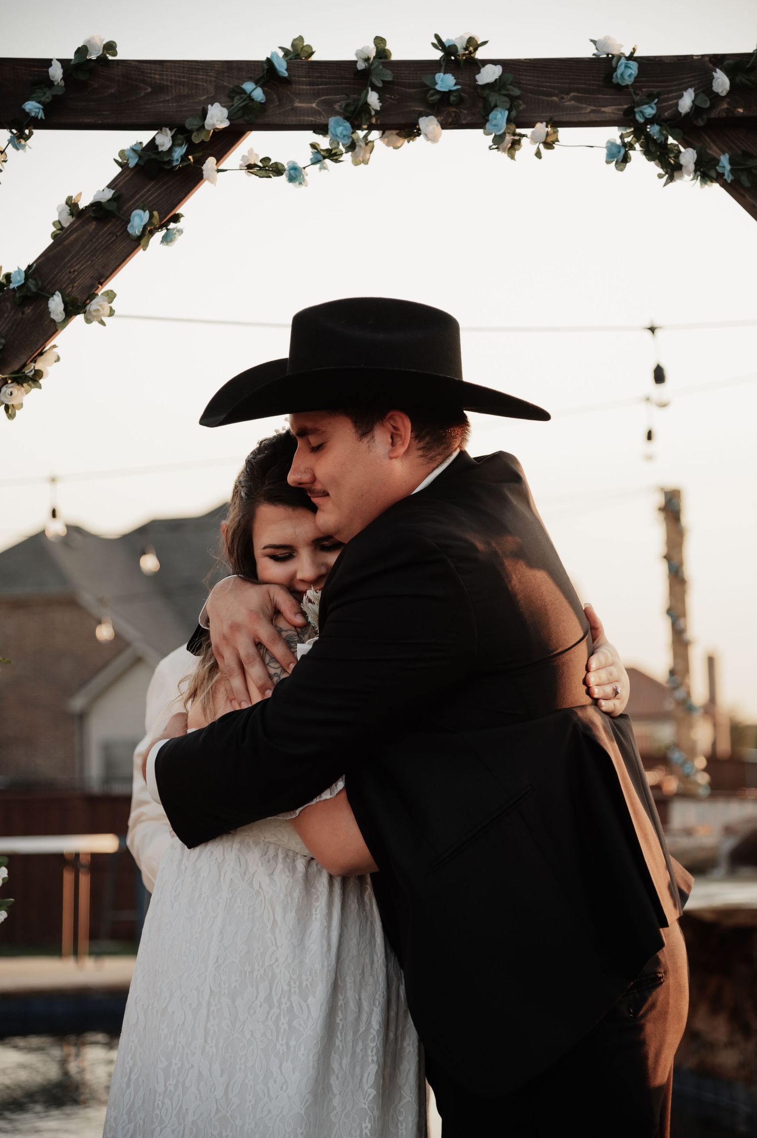 Bride and groom share a heartfelt hug under a floral arch at an outdoor wedding ceremony at dusk.