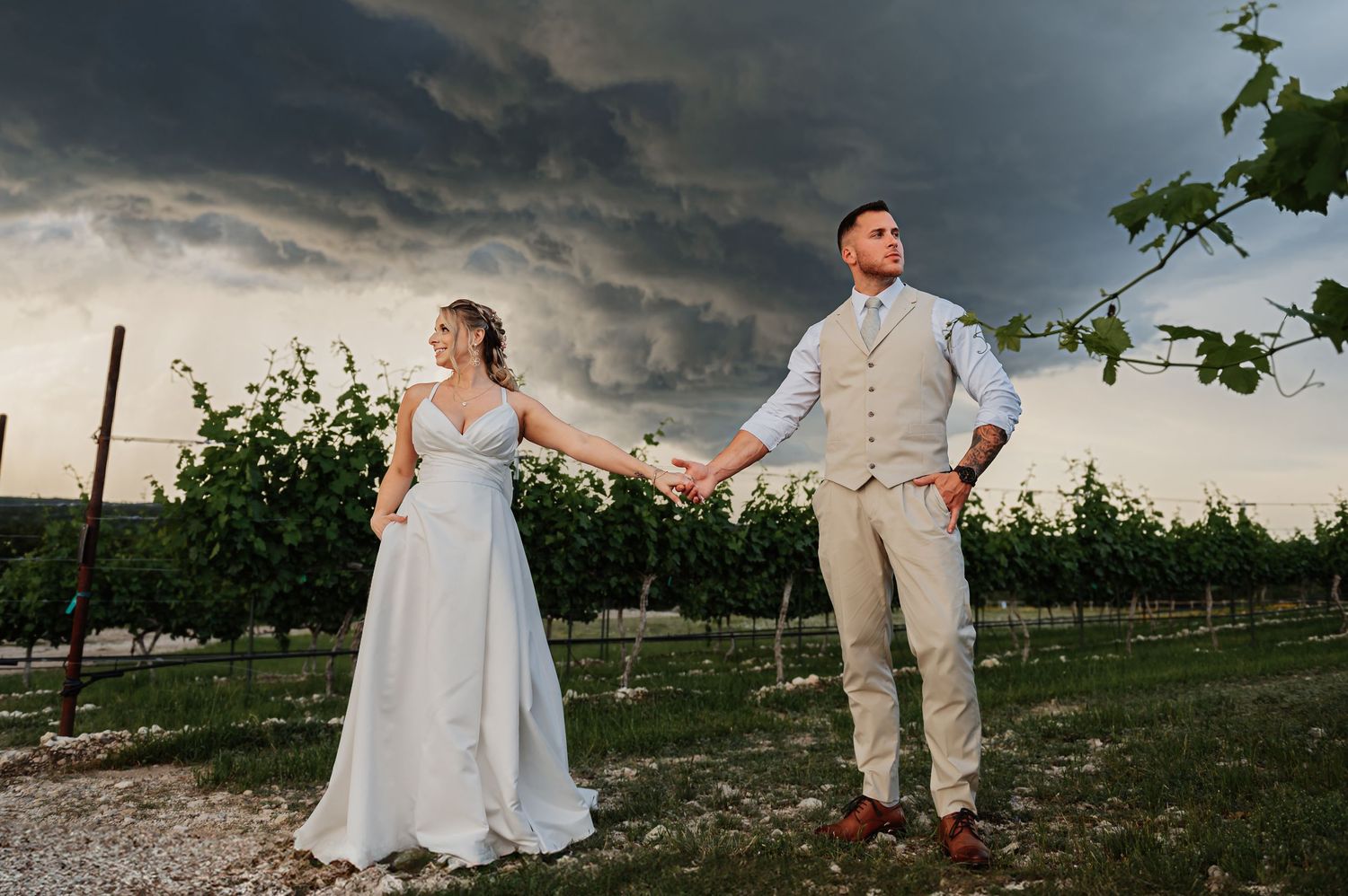 Bride and groom hold hands in a vineyard under dramatic storm clouds on their wedding day.
