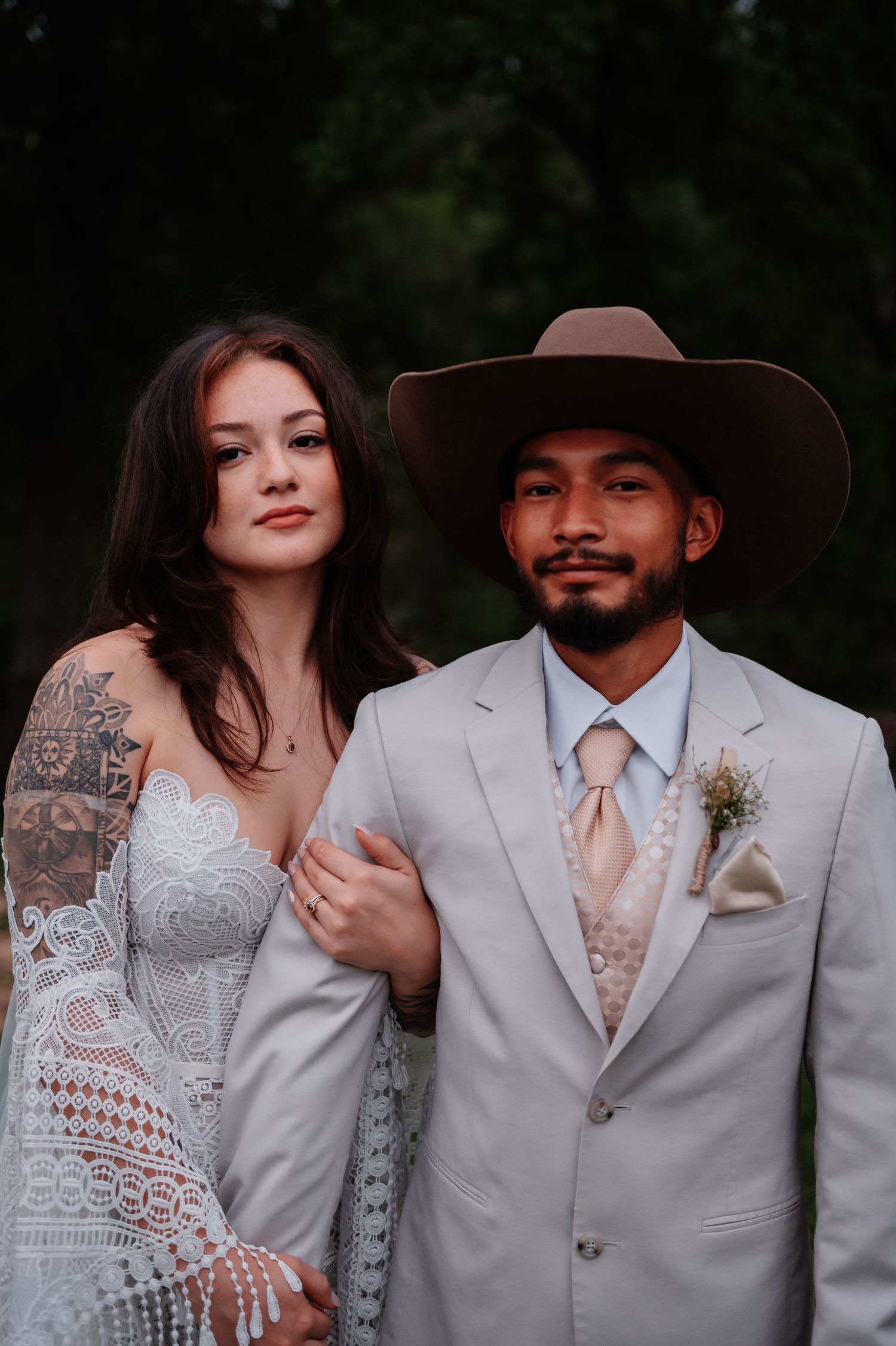 A bride in lace wedding dress with tattoos stands beside groom in gray suit and cowboy hat with boutonniere.