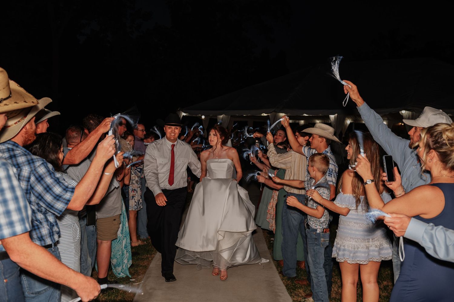 Bride and groom exit wedding ceremony through cheering guests waving items at night outdoor celebration.