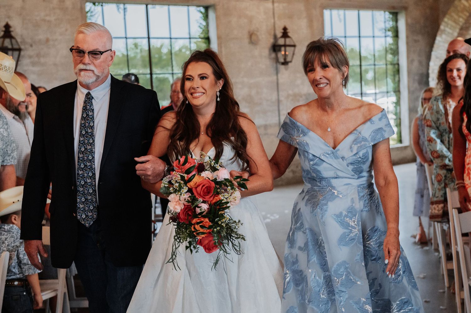 Bride walks down the aisle with parents at elegant indoor wedding ceremony, holding vibrant red and orange bouquet.