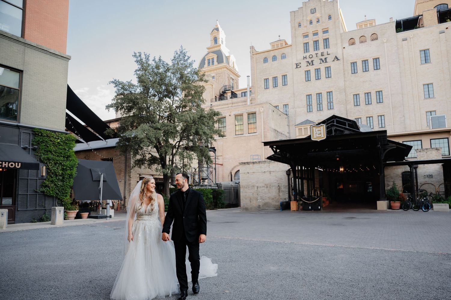 Bride and groom stand in a sunlit courtyard with a grand white historic building backdrop at golden hour.