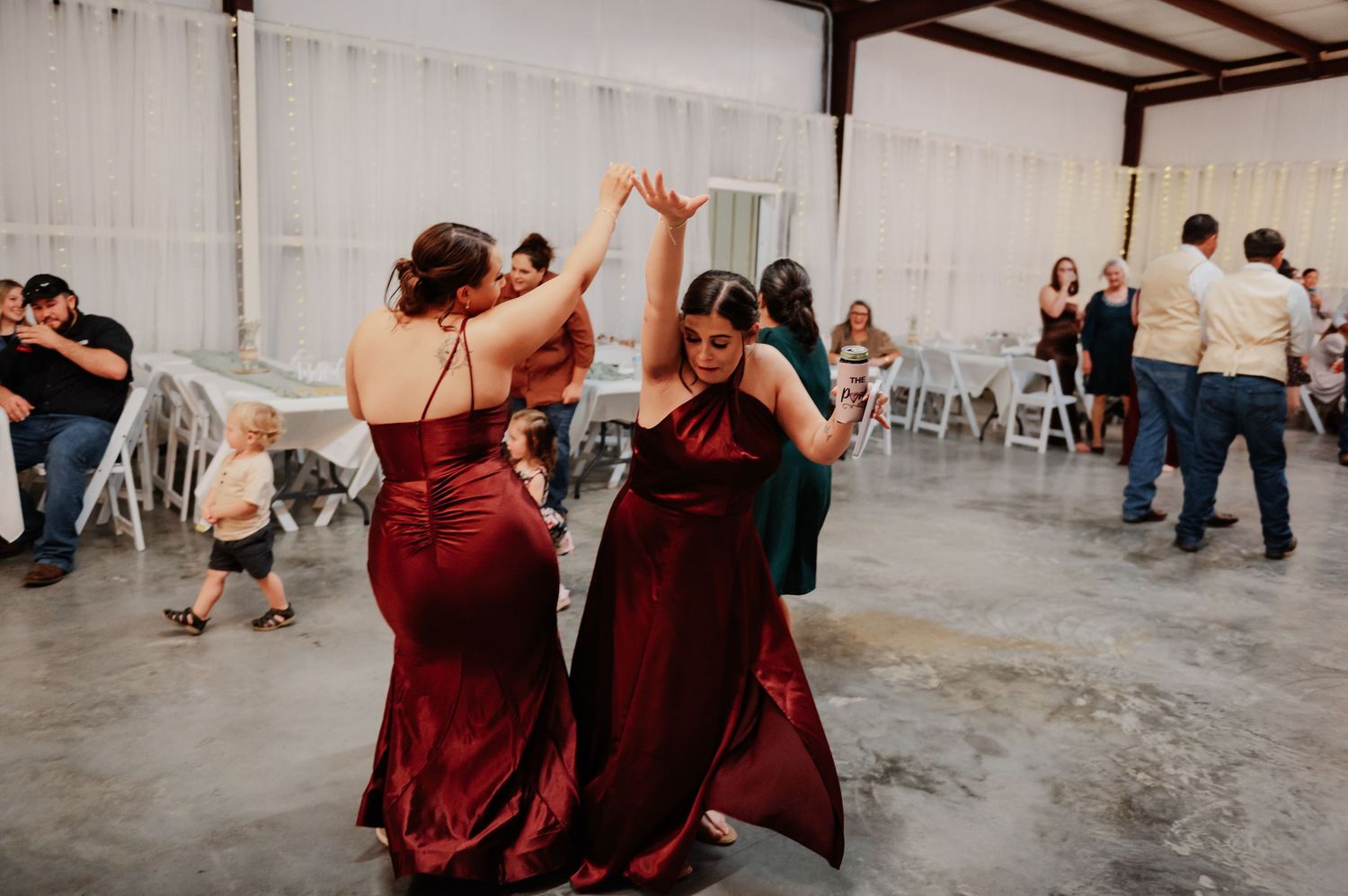 Two bridesmaids in burgundy satin gowns dancing joyfully at an indoor wedding reception venue.