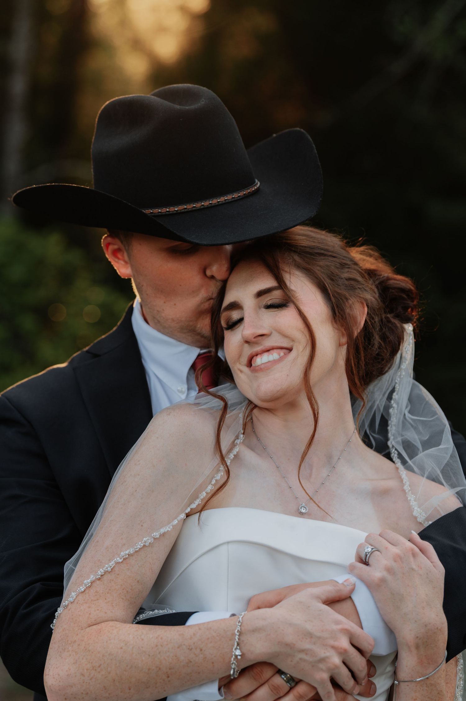 Groom in black cowboy hat kissing bride's cheek as she smiles, wearing white dress and veil at outdoor wedding.
