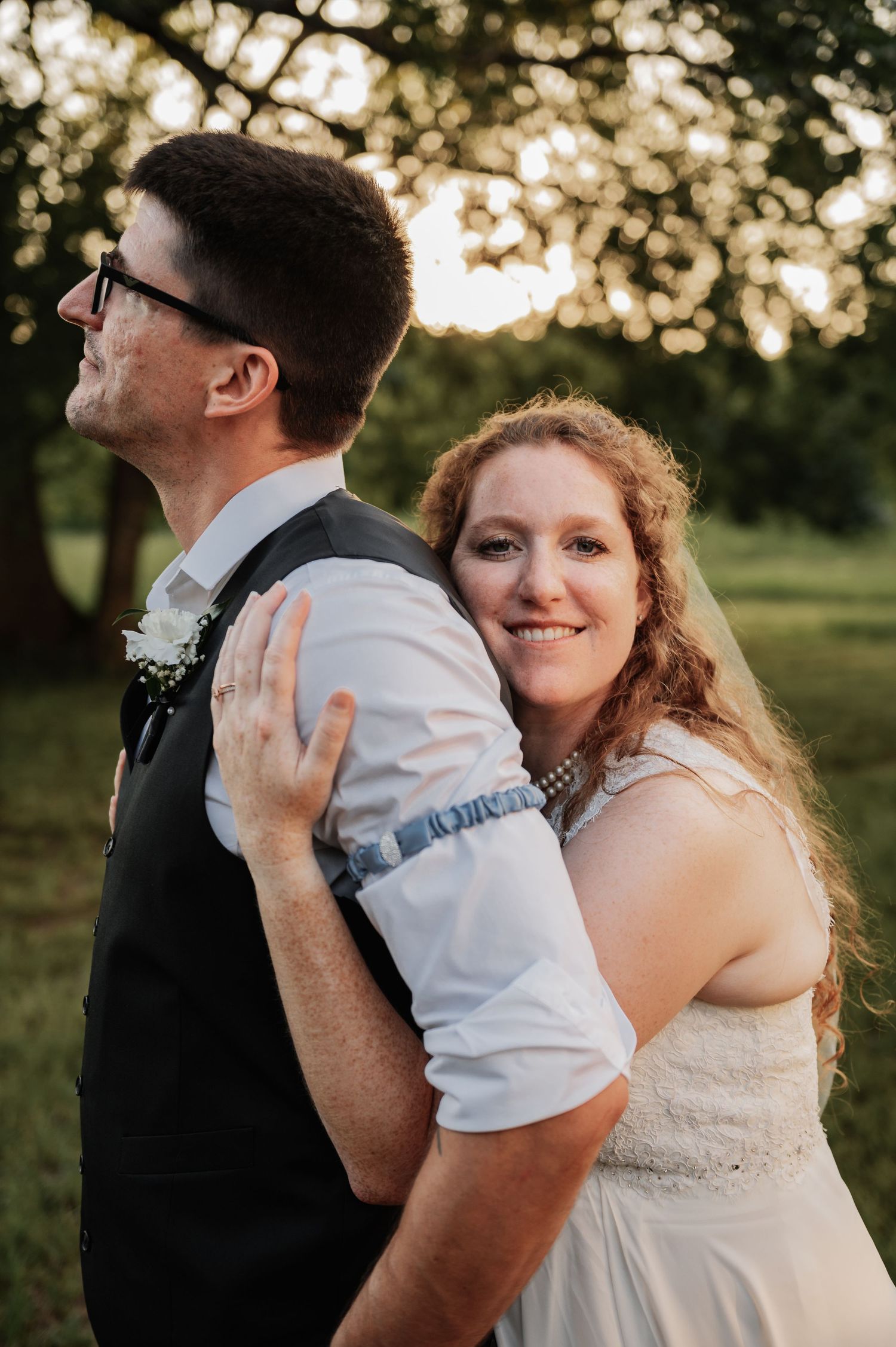 A bride with red curly hair embraces a groom in a black vest outdoors during golden hour at their wedding.
