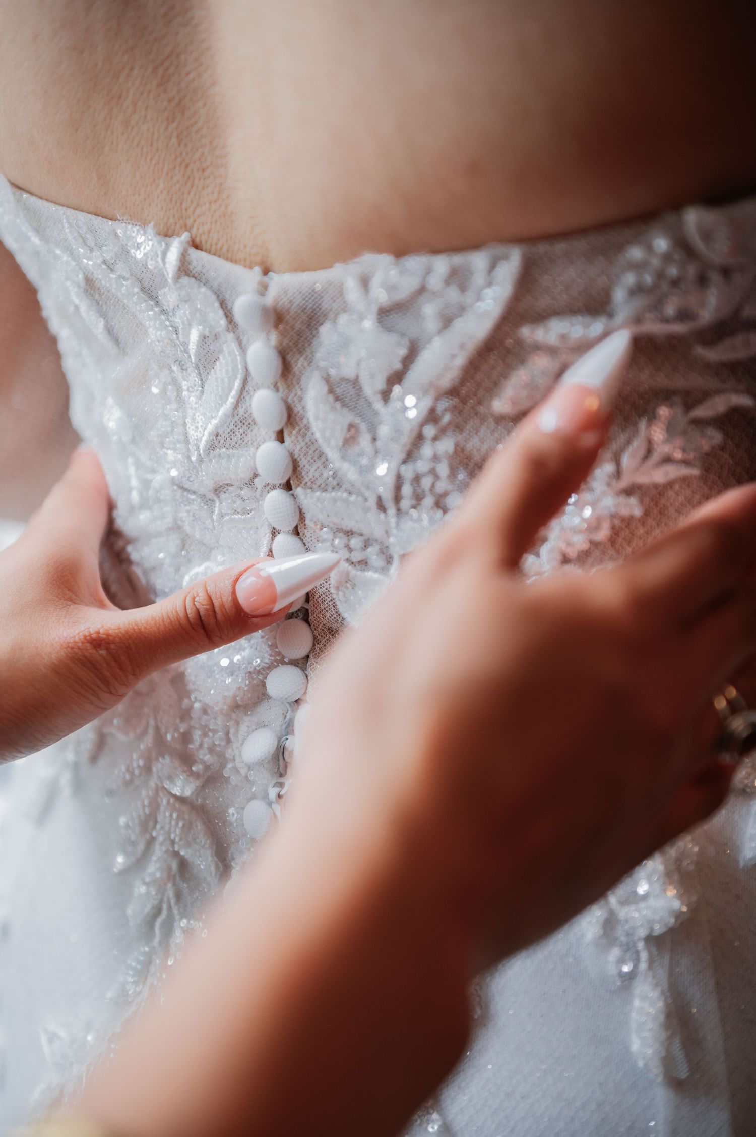 Hands fastening delicate white buttons on the back of an elegant lace wedding dress.