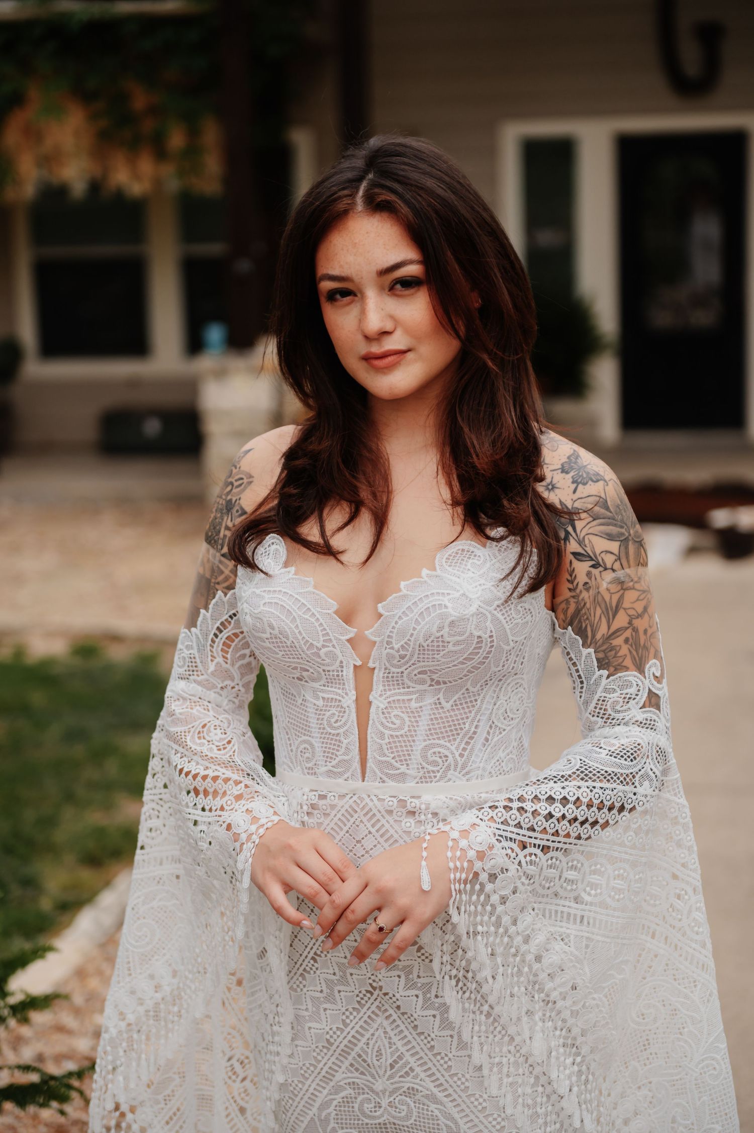 Bride in white lace bell-sleeve wedding gown with tattoos, posing outdoors at a venue.