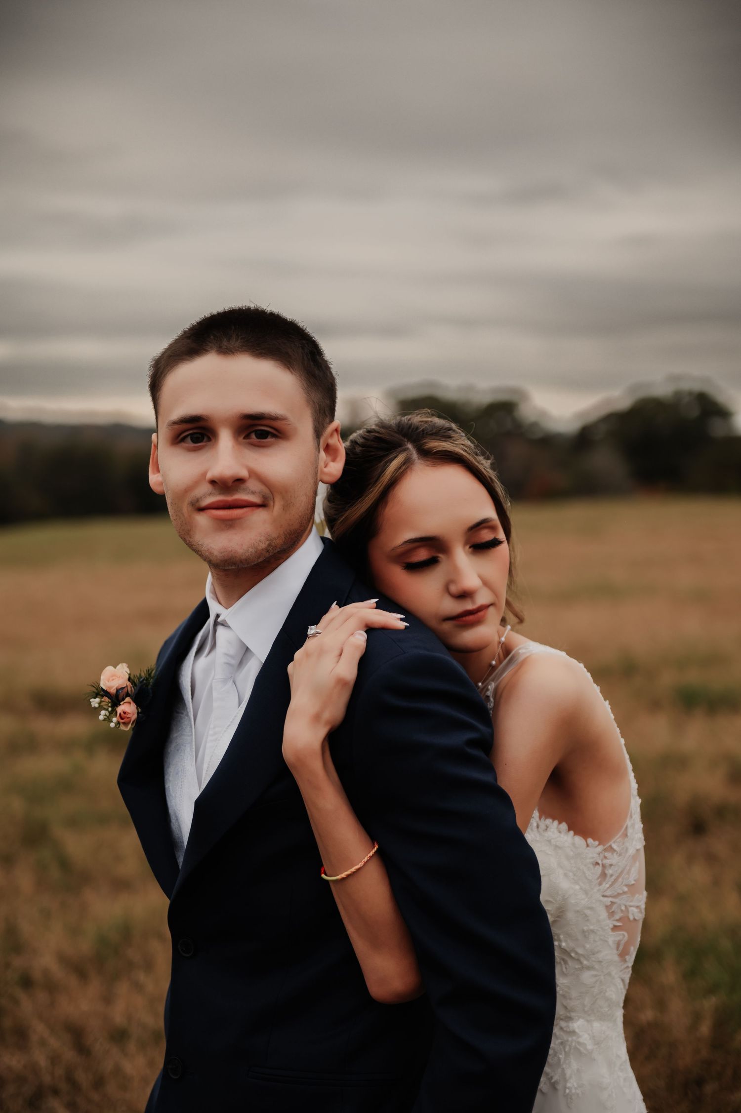 A bride in lace gown embraces a groom in navy suit with boutonniere in an autumn field under overcast skies.