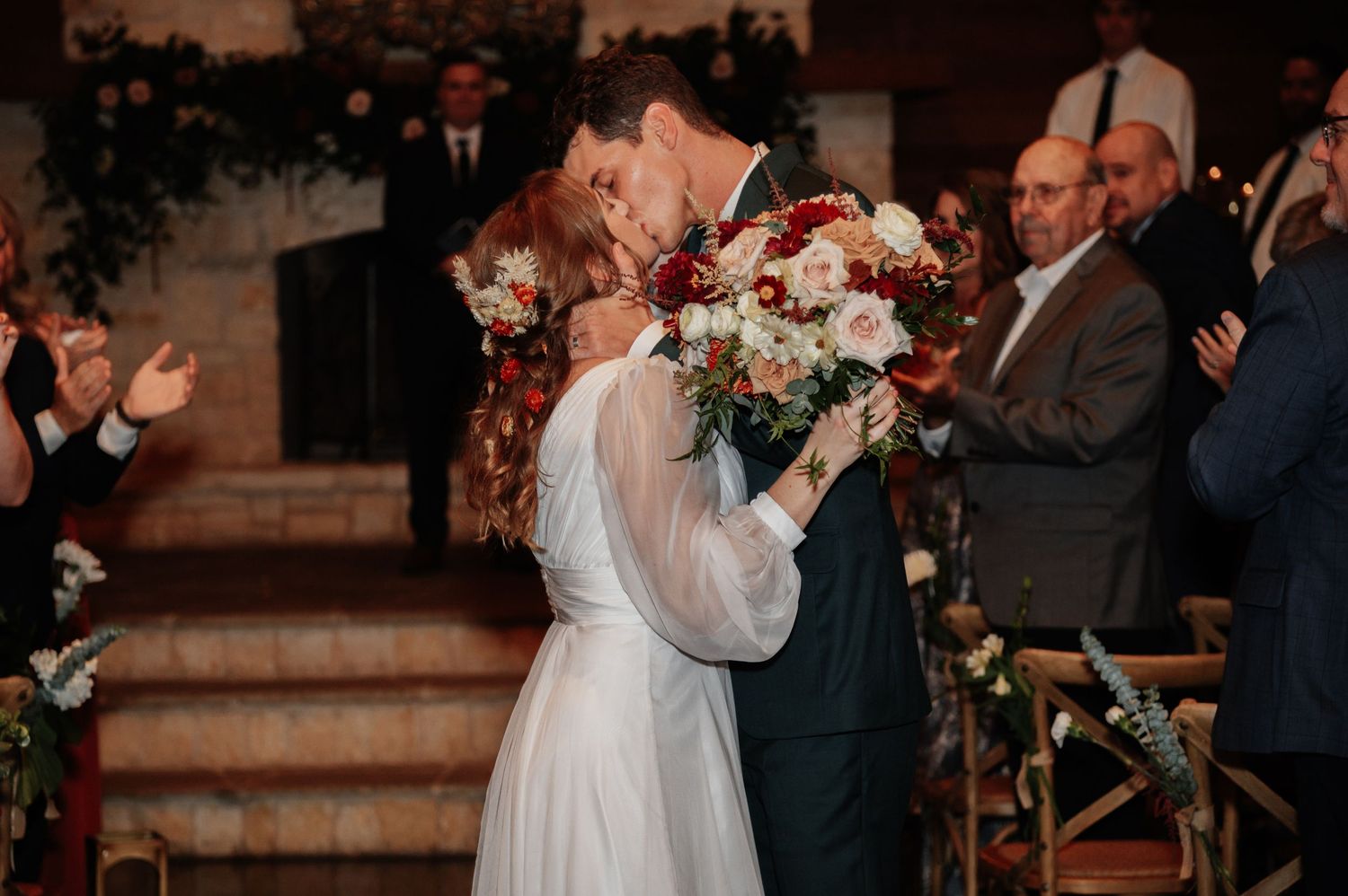 Bride and groom share first kiss at wedding altar, bride holding red and white bouquet with guests applauding.