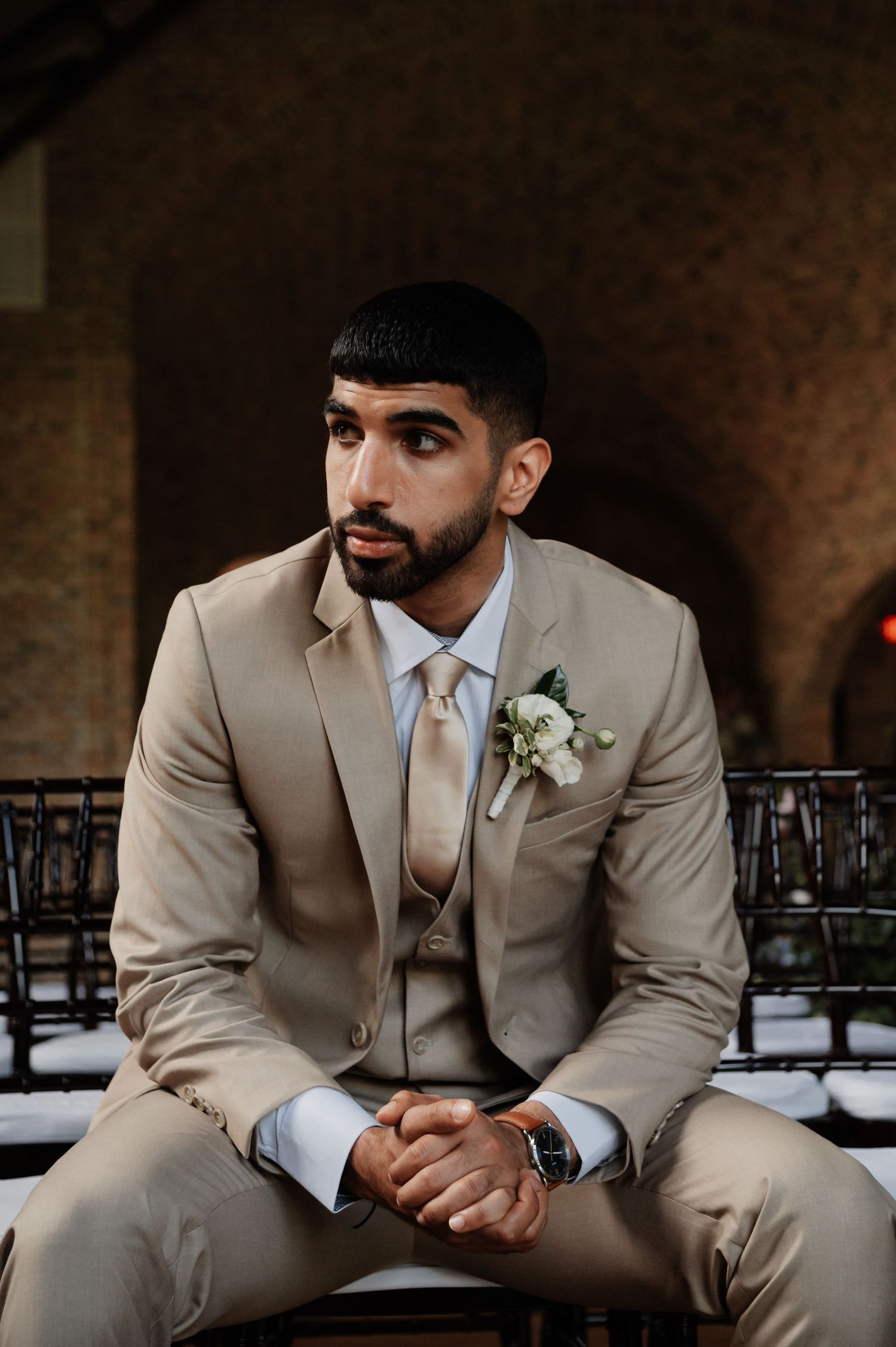 Groom in tan three-piece suit with boutonniere sits thoughtfully at wedding ceremony venue.