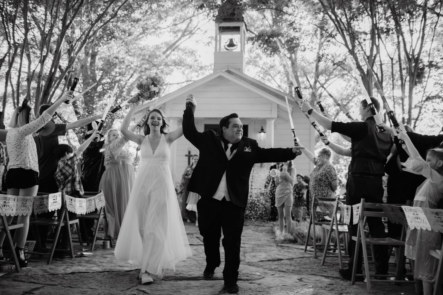 Bride and groom celebrate outdoor wedding exit with sparklers near a charming white chapel in black and white photo.