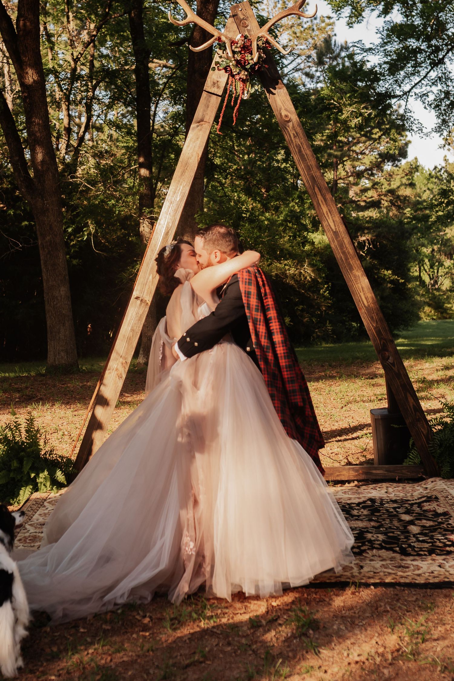 Bride and groom share first kiss under rustic wooden arch in sunlit forest setting, groom wearing red plaid jacket.