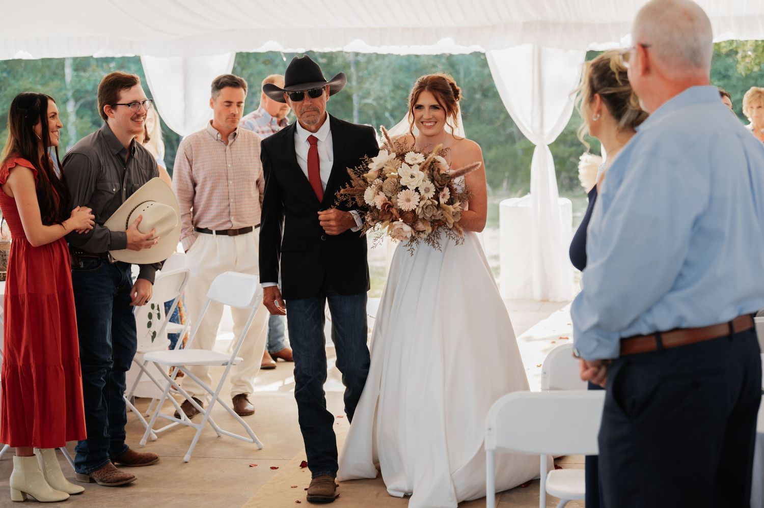 Bride walks down the aisle with her father at an outdoor wedding ceremony under a white tent.