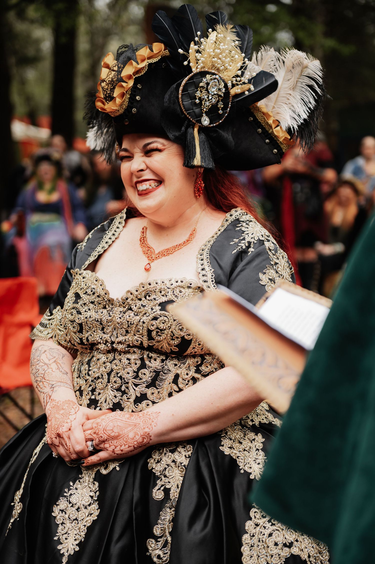 Woman in black and gold Renaissance gown with feathered hat laughs joyfully at outdoor costume festival.
