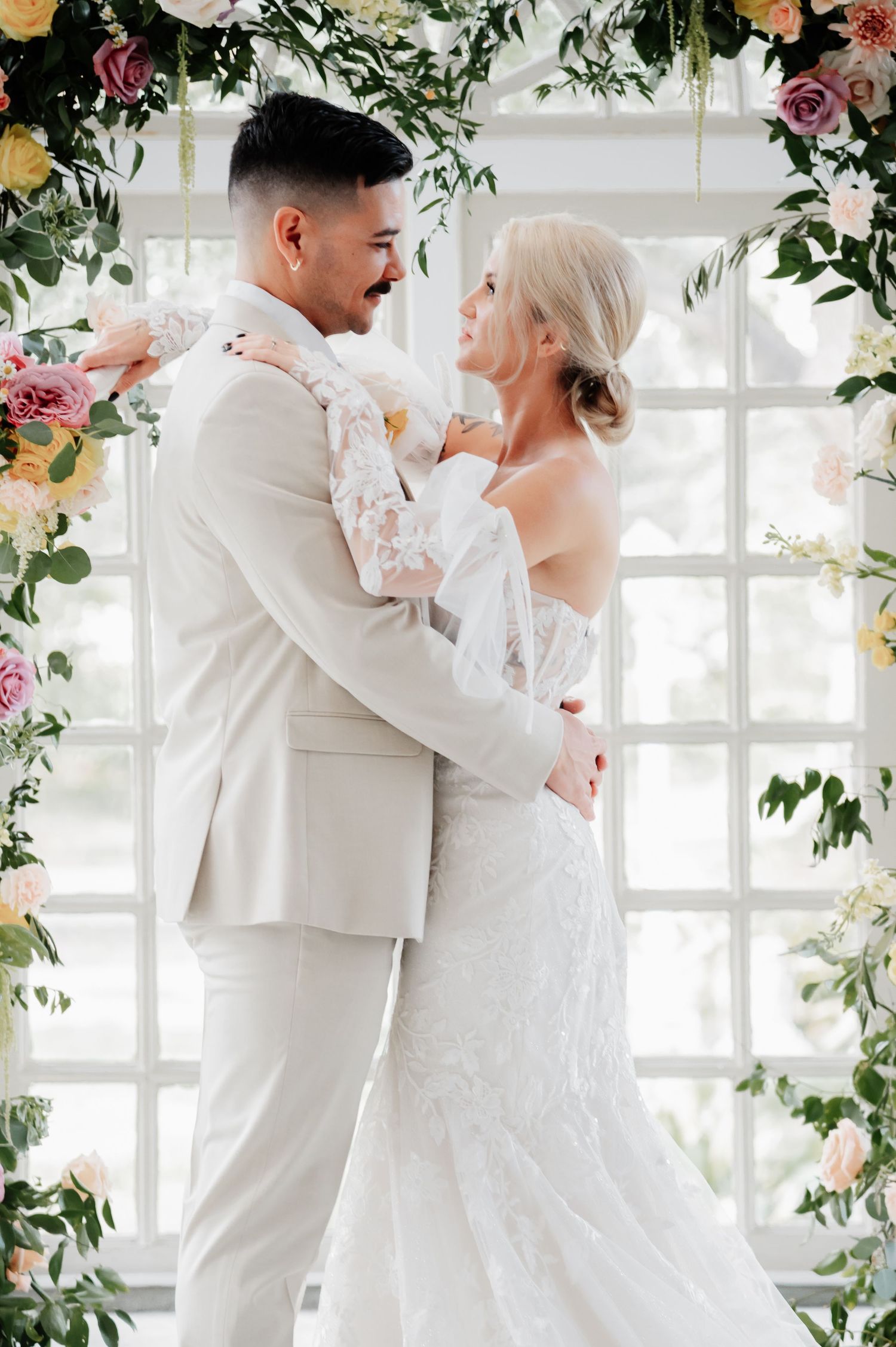 Bride and groom embracing under floral arch with pink and yellow blooms, sharing a romantic moment at their wedding ceremony.