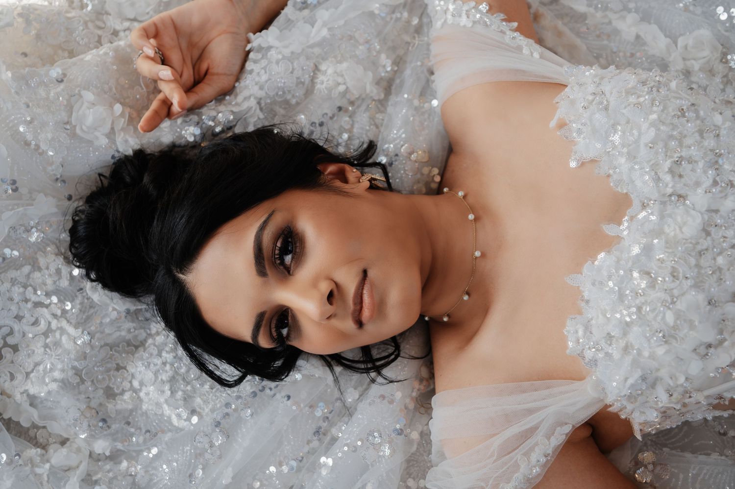 Woman in elegant white lace bridal gown lying down, wearing pearl necklace with dramatic dark makeup.