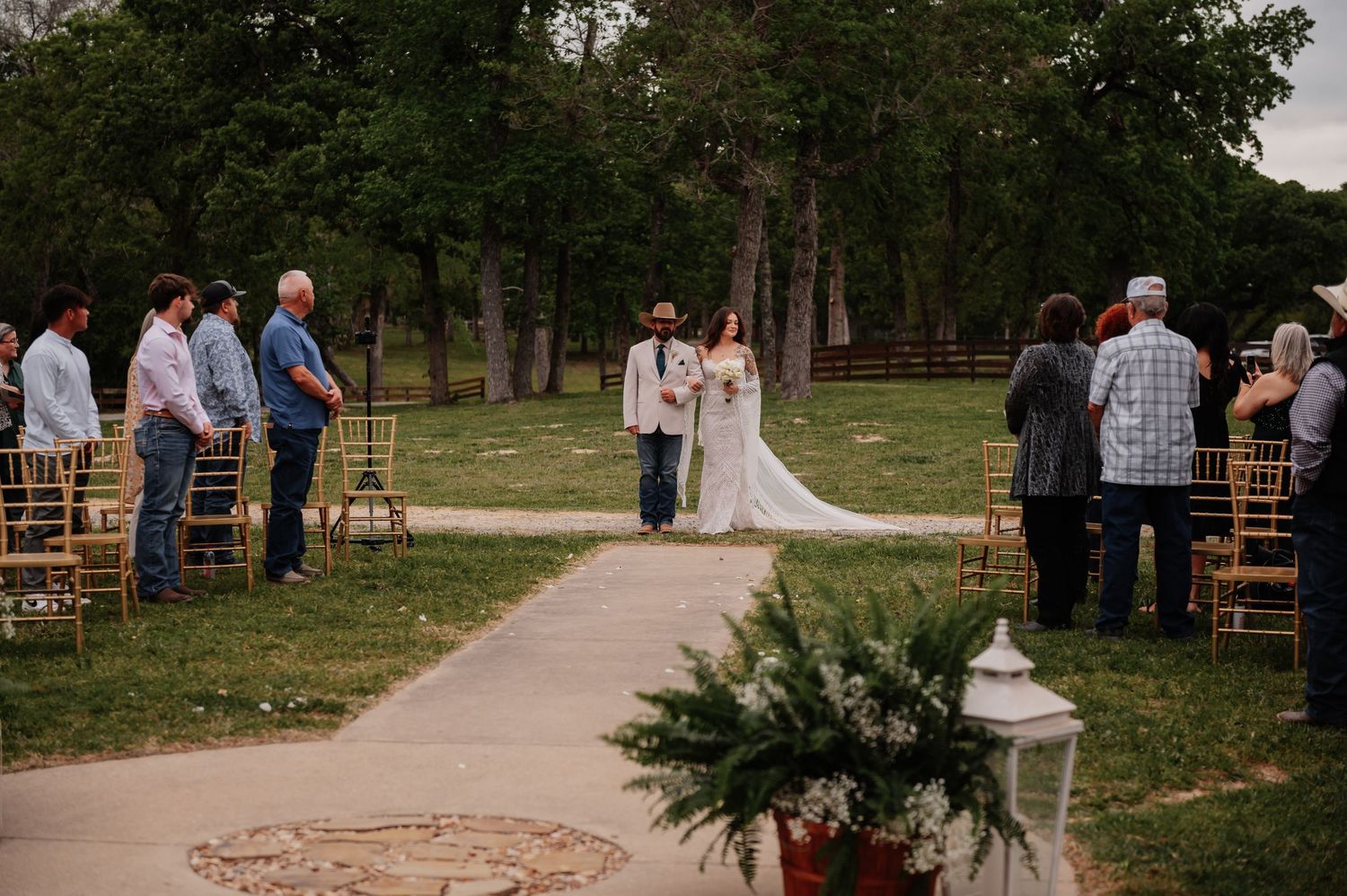 Outdoor wedding ceremony with guests standing along a rustic aisle, bride and groom at the altar surrounded by trees.