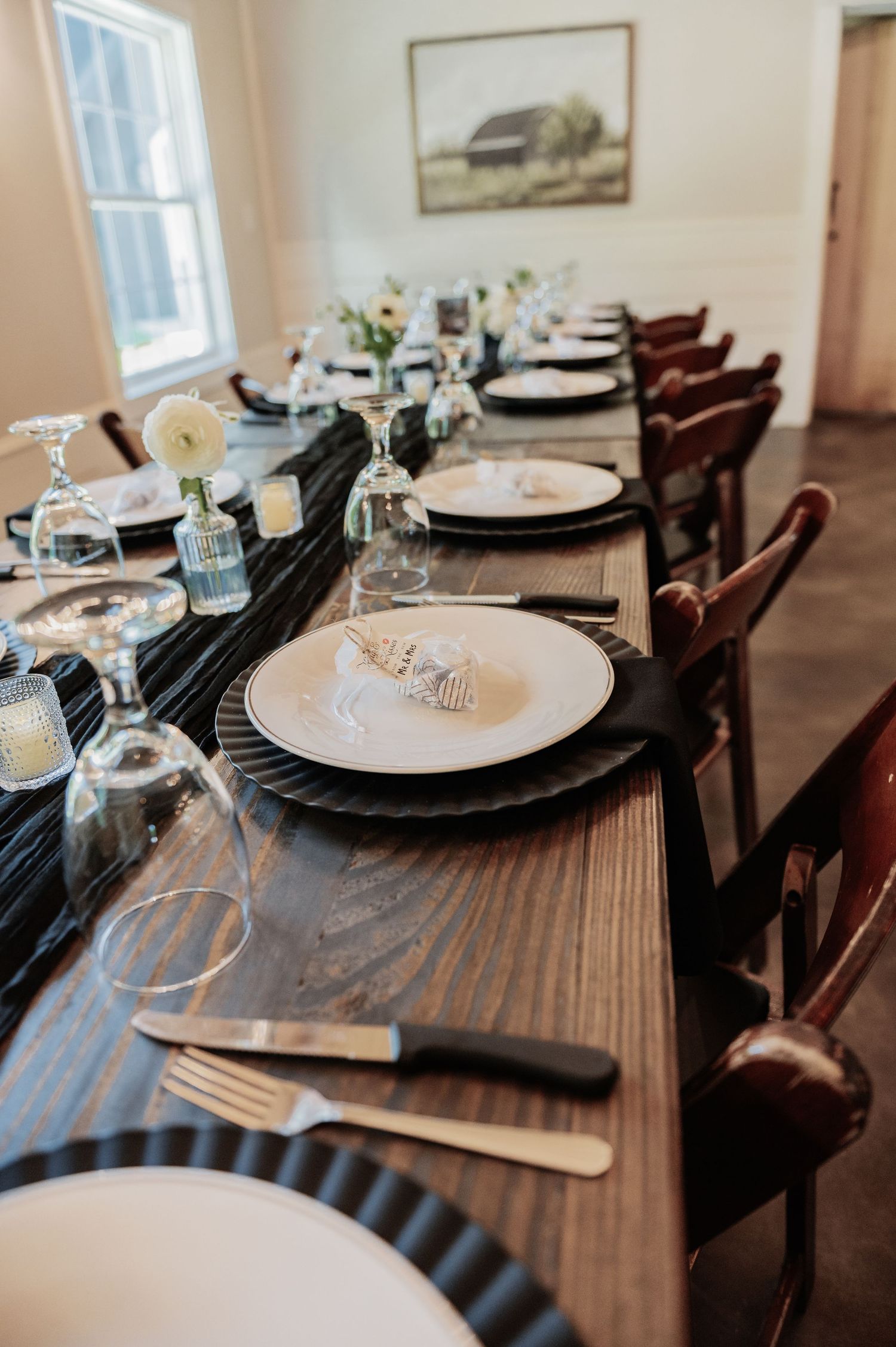 Elegant farmhouse dining table set with black chargers, white plates, gold cutlery, and floral centerpieces for a formal event.