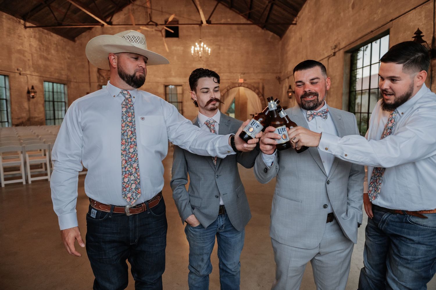 Four groomsmen in gray suits toasting with beer bottles in a rustic wedding venue.