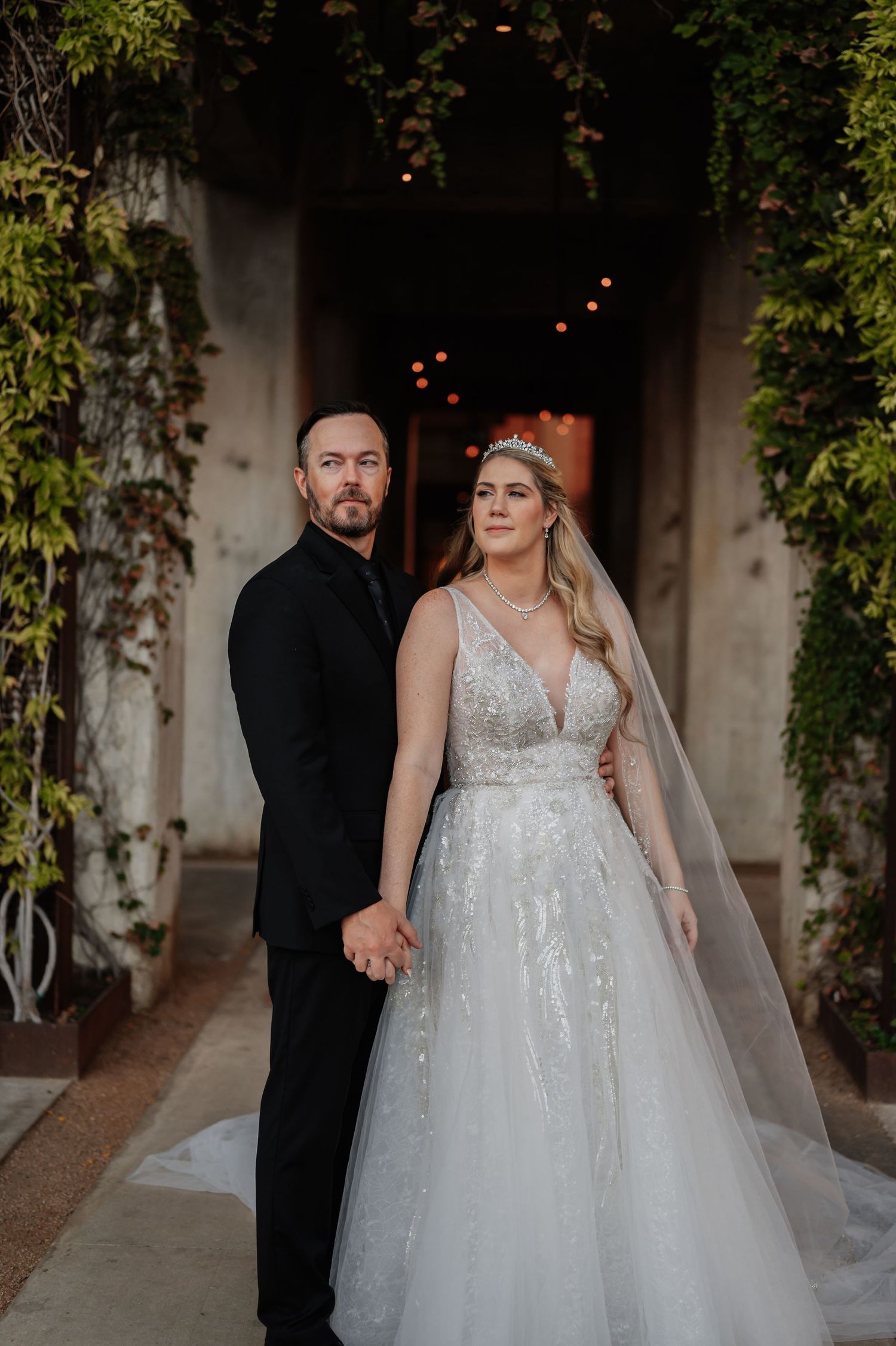 Bride in beaded gown with veil and groom in black suit hold hands at elegant ivy-covered wedding venue.