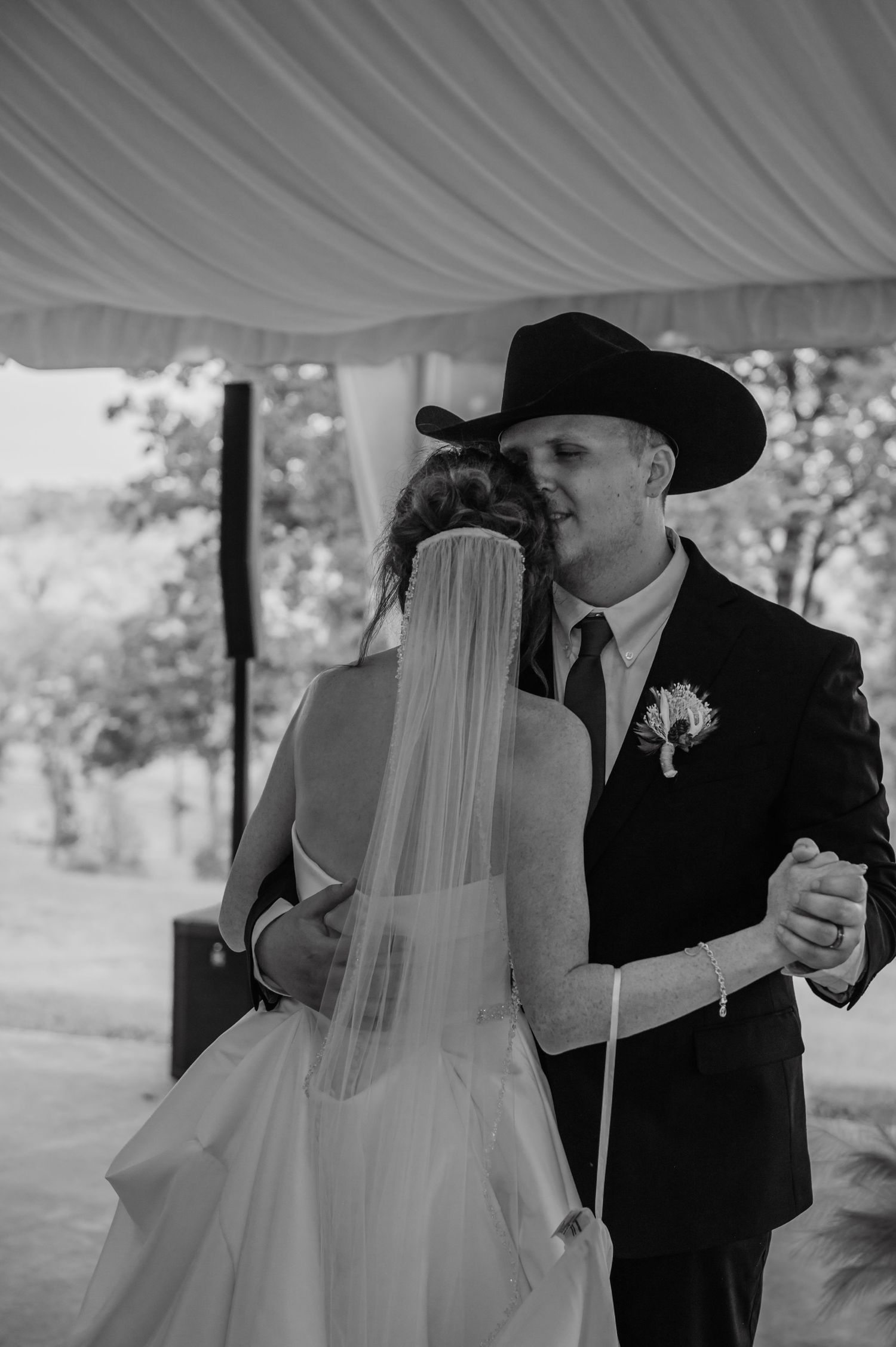 Black and white photo of a bride and groom sharing their first dance, groom wearing a cowboy hat and boutonniere.