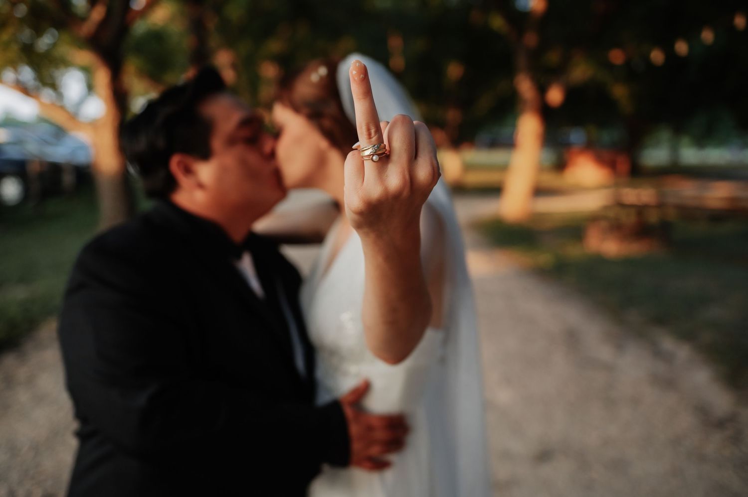 Bride shows off wedding ring while kissing groom outdoors at golden hour in a romantic park setting.