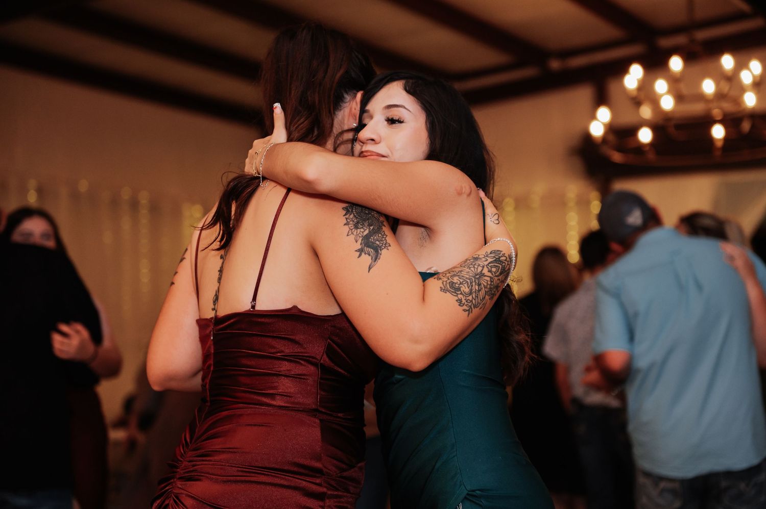 Two women in elegant gowns share a warm embrace on a wedding reception dance floor.