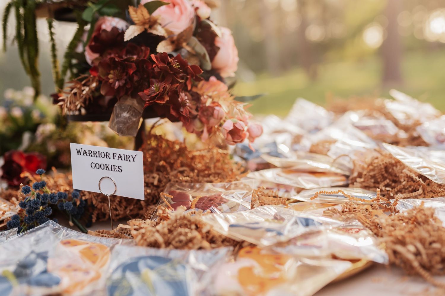 Rustic wedding dessert table with warrior fairy cookies in cellophane bags surrounded by dried florals and fresh flower arrangement.