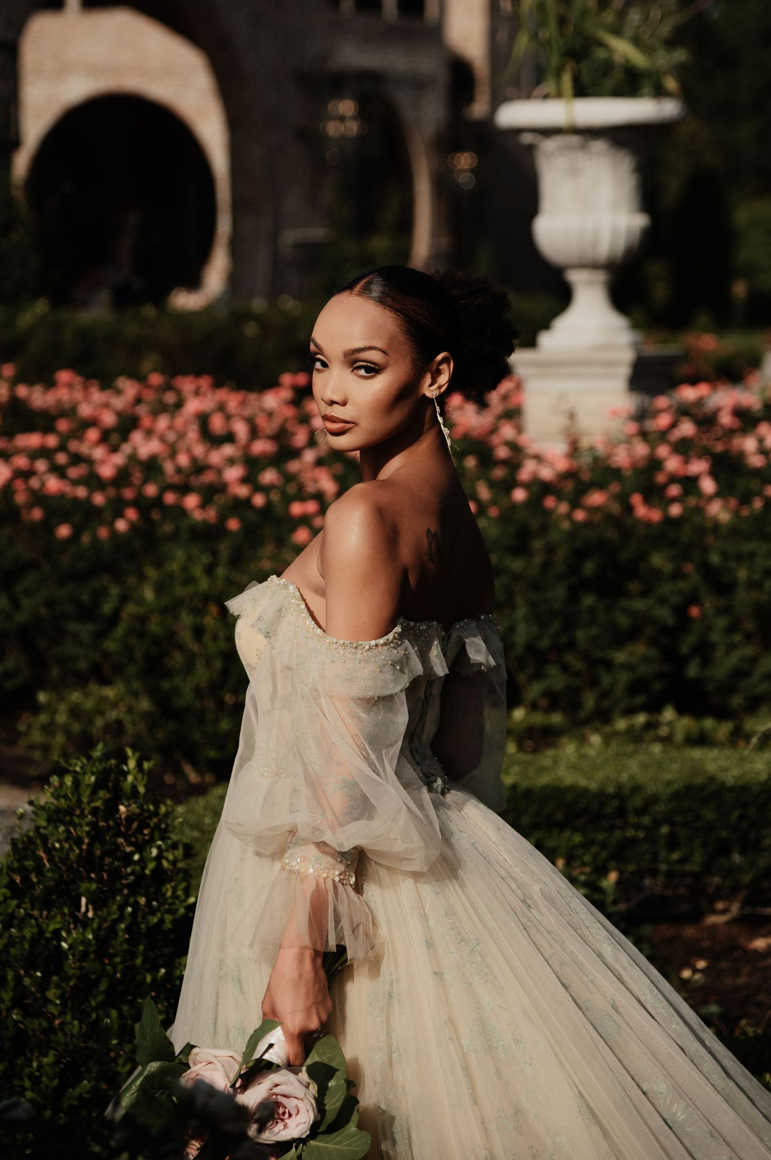 Bride in a sheer off-shoulder tulle gown poses in a garden surrounded by pink roses and lush greenery.