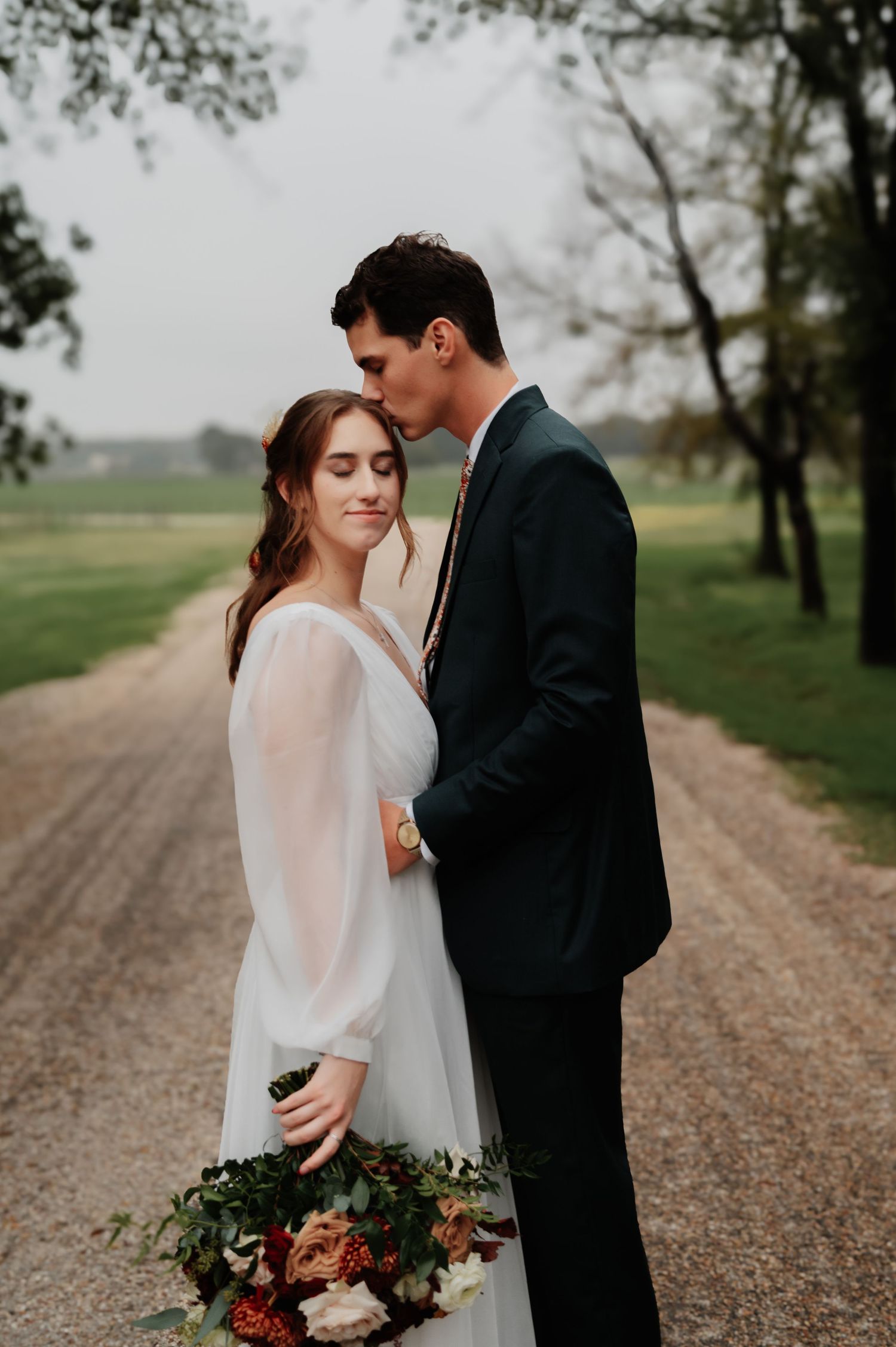Bride in a flowing white gown and groom in a dark suit share a romantic moment on a tree-lined gravel path.