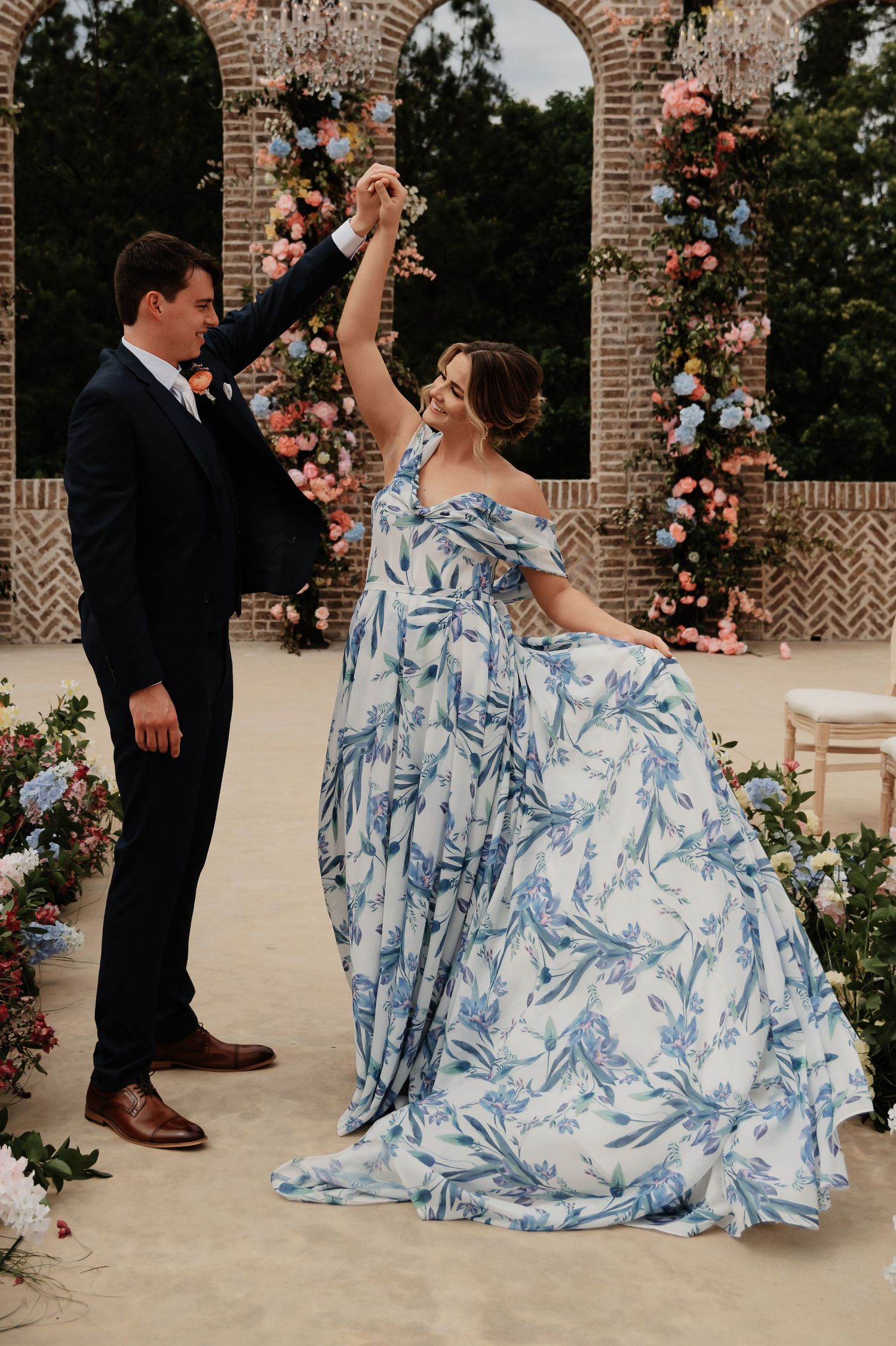 Bride in blue floral off-shoulder gown twirls with groom in navy suit at outdoor wedding ceremony.