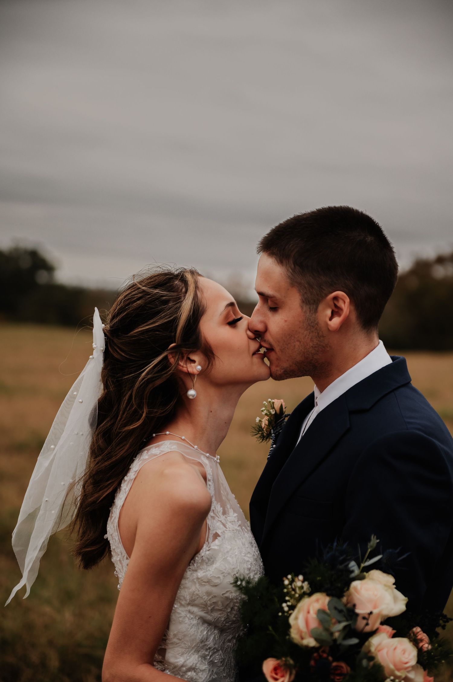 A bride and groom share a romantic kiss outdoors in a field, with the bride holding a bouquet and wearing a lace gown.