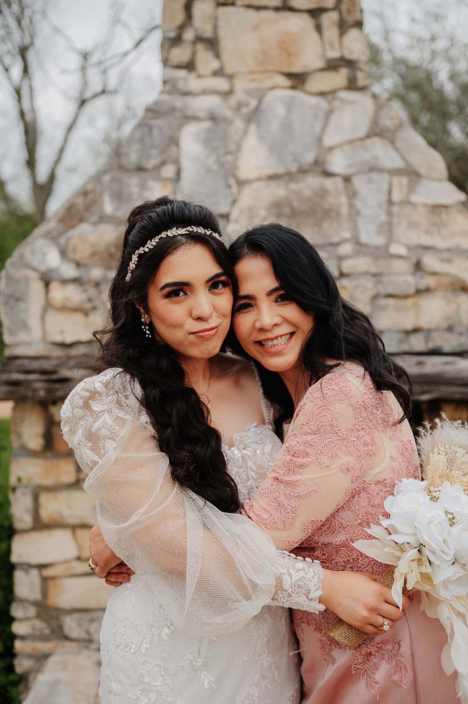 Bride in white lace gown embraces woman in pink dress holding white bouquet before stone wall.