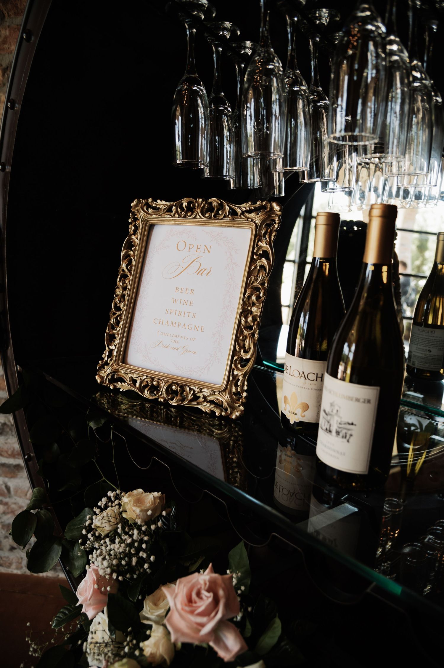 Elegant bar setup with gold-framed open bar sign, white wine bottles, and hanging champagne glasses at a wedding reception.