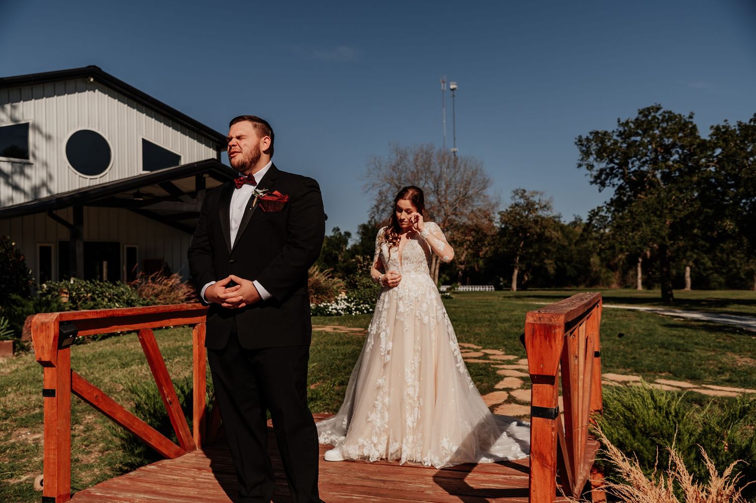 Bride in lace gown and groom in black tuxedo share an emotional first look on a wooden deck outdoors.
