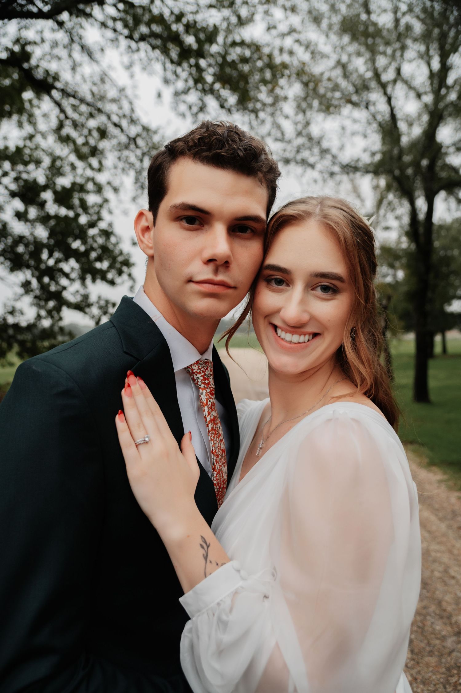 A smiling bride in a white dress and groom in a dark suit with floral tie pose together outdoors among trees.