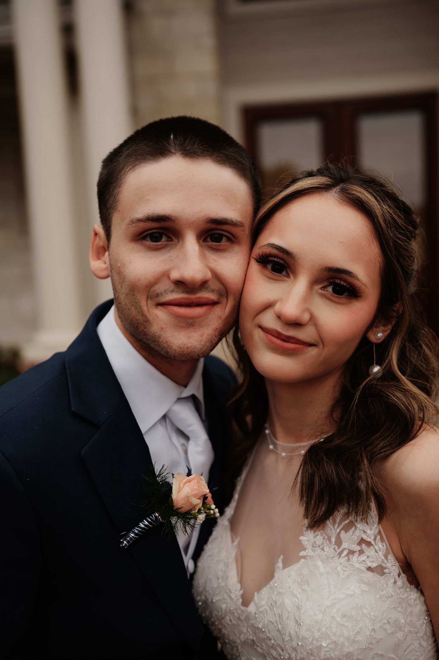 A smiling couple in wedding attire, groom in navy suit with boutonniere, bride in lace dress, posing cheek to cheek outdoors.
