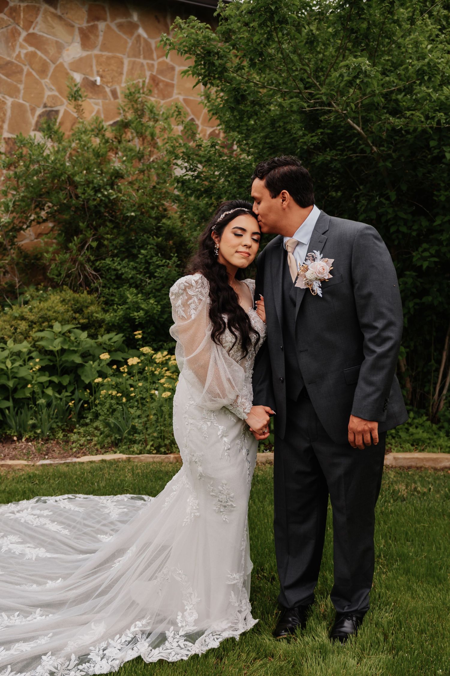 Bride in lace gown with train and groom in gray suit share a tender kiss in a lush garden setting.