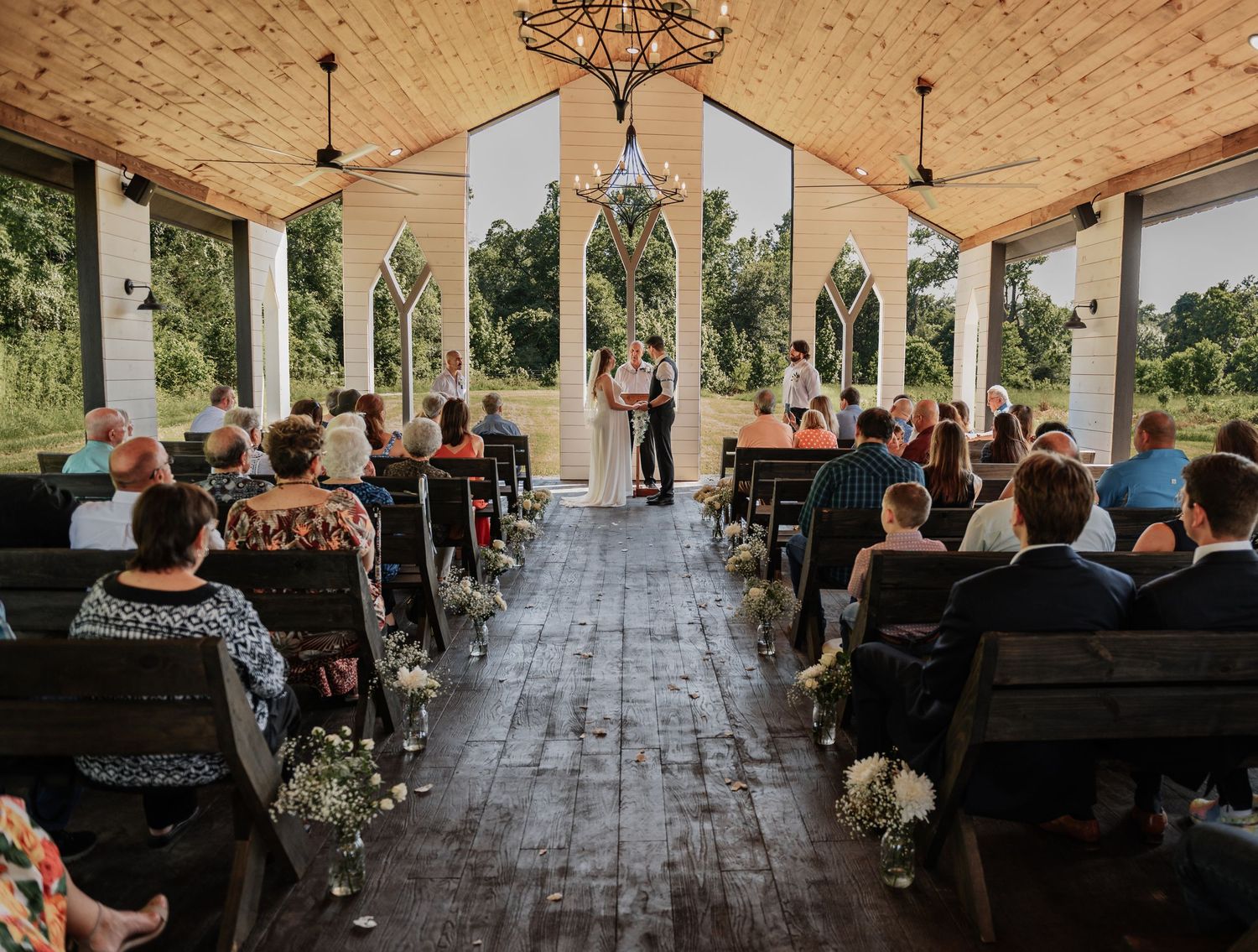 Rustic outdoor wedding ceremony in an open-air chapel with wooden pews, chandelier, and lush green surroundings.