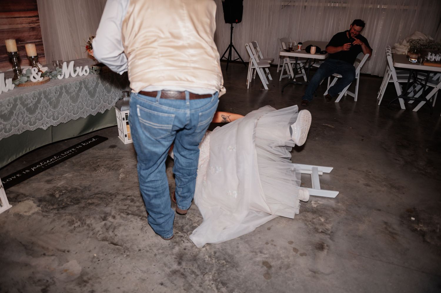 Person in jeans assists a bride in a white wedding gown at an elegant indoor wedding reception venue.