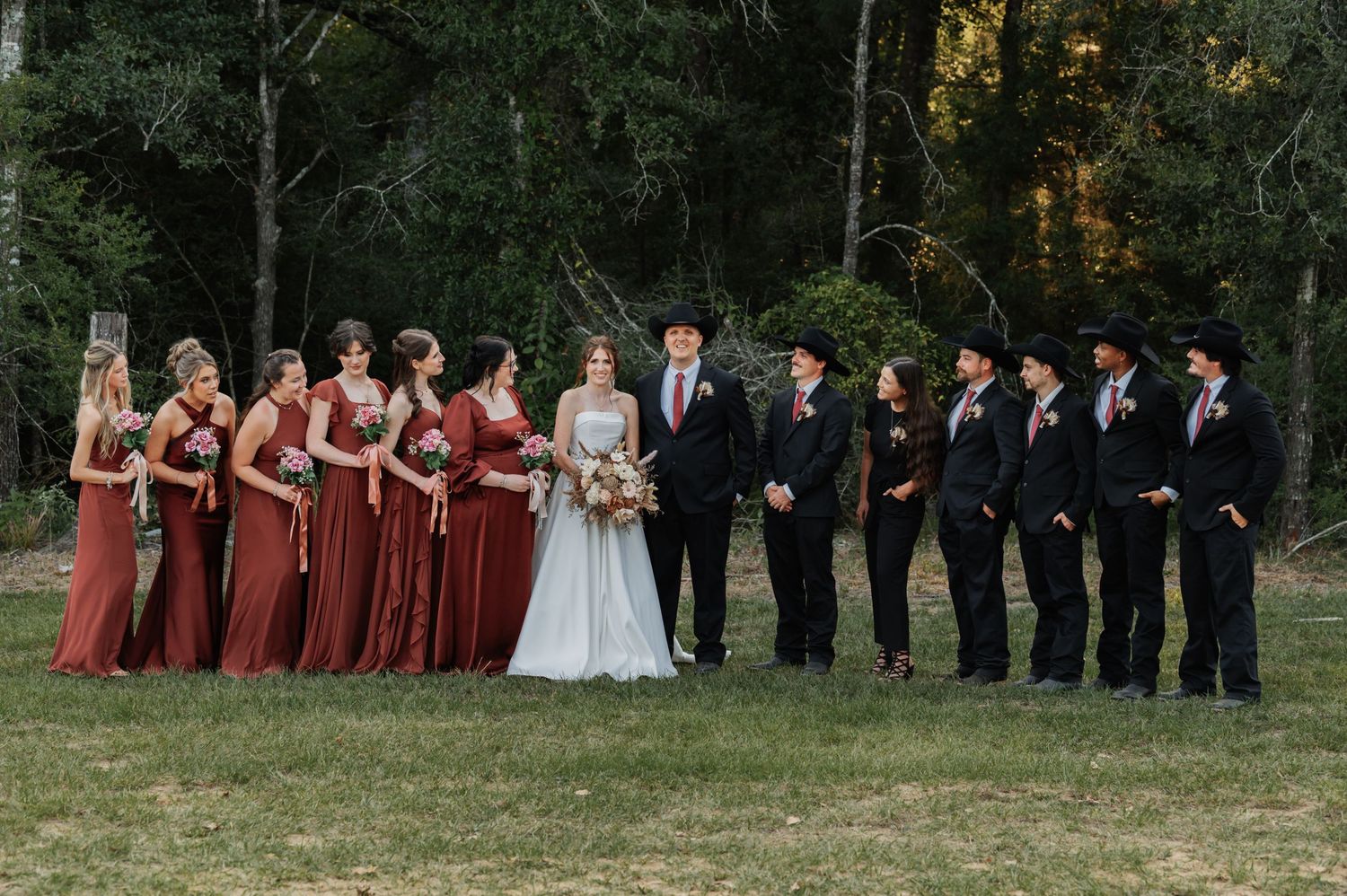 Wedding party poses outdoors with bridesmaids in red dresses and groomsmen in black suits with cowboy hats.