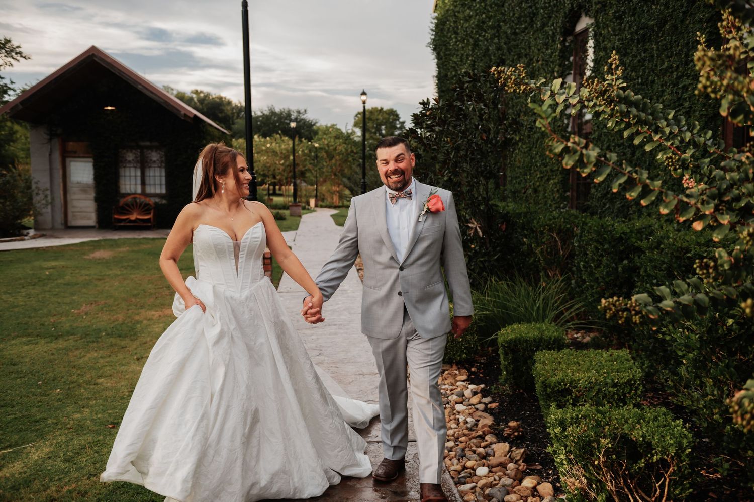 Bride and groom walk hand in hand through lush garden path near rustic venue on their wedding day.