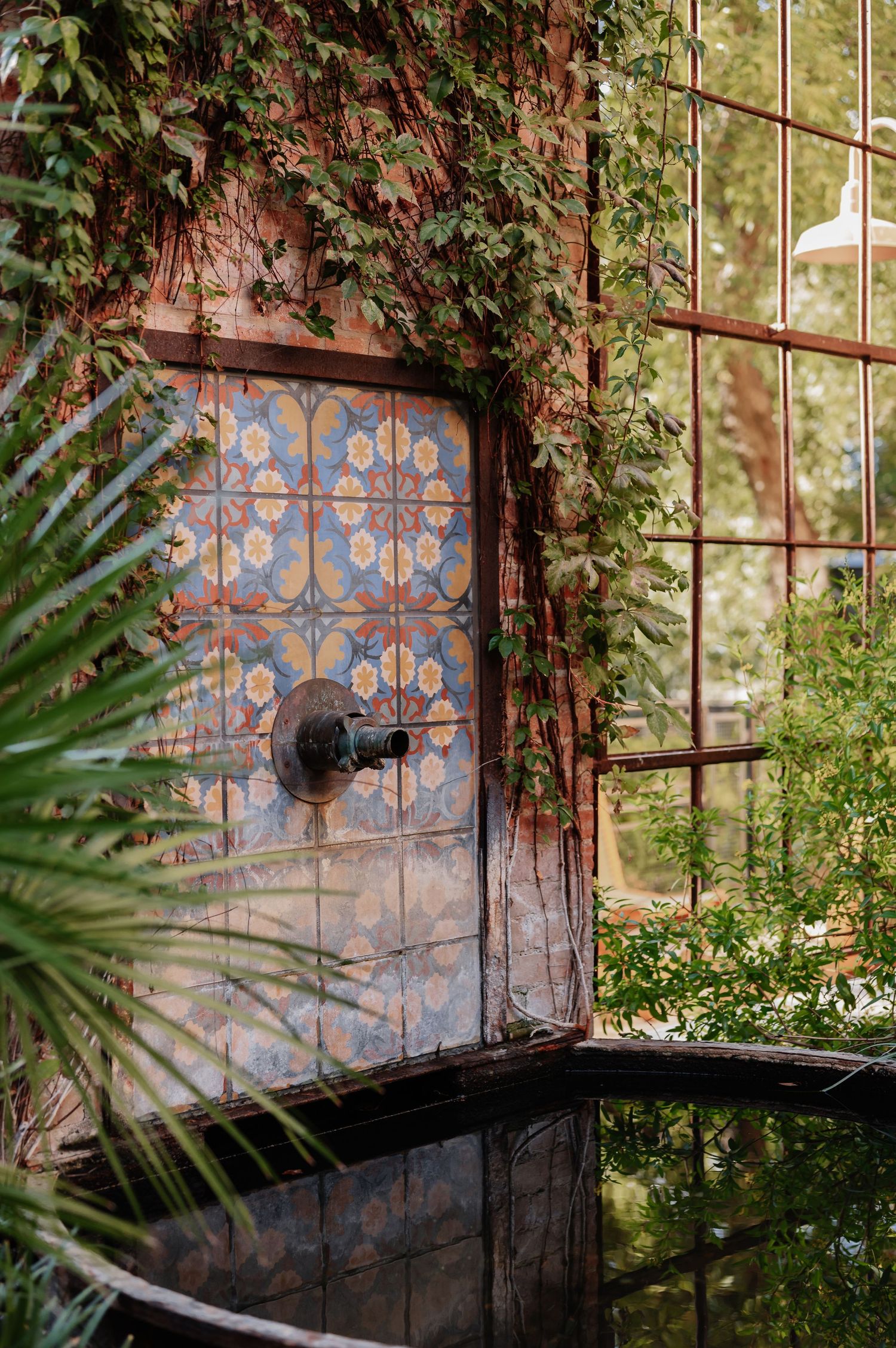Rustic tiled wall fountain with blue mosaic patterns, draped in lush green ivy, surrounded by tropical plants in a garden setting.