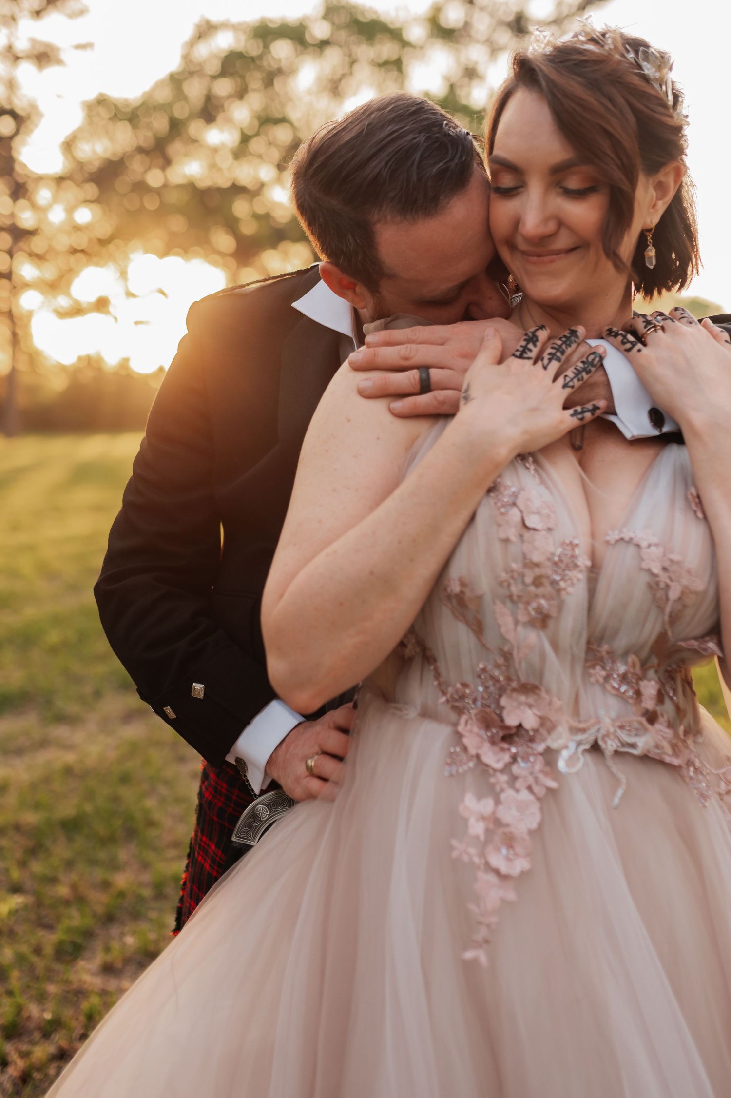 Groom in Scottish kilt kissing bride in blush floral gown at golden hour outdoor wedding ceremony.