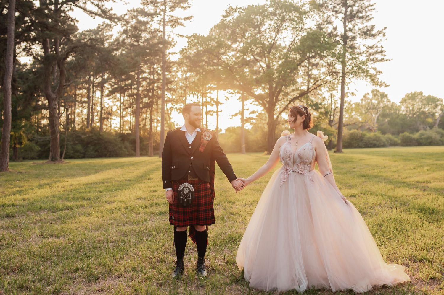 Bride and groom holding hands at sunset in a field, groom wearing a red tartan kilt and bride in a blush ball gown.
