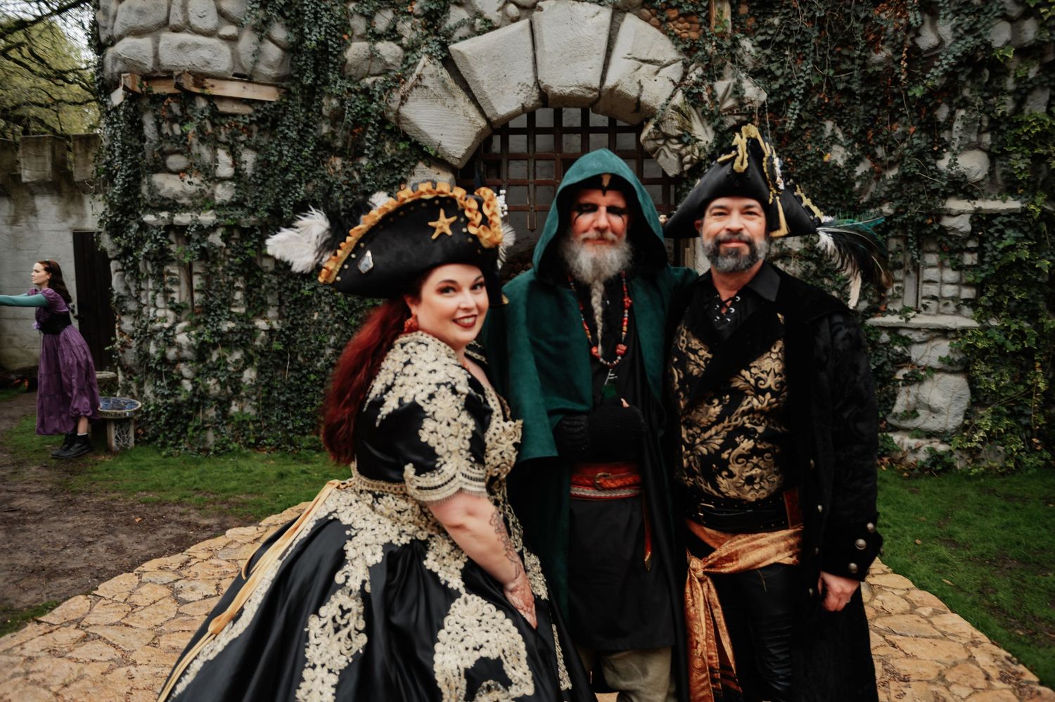 Three people in elaborate Renaissance faire costumes pose together outdoors near a stone wall.