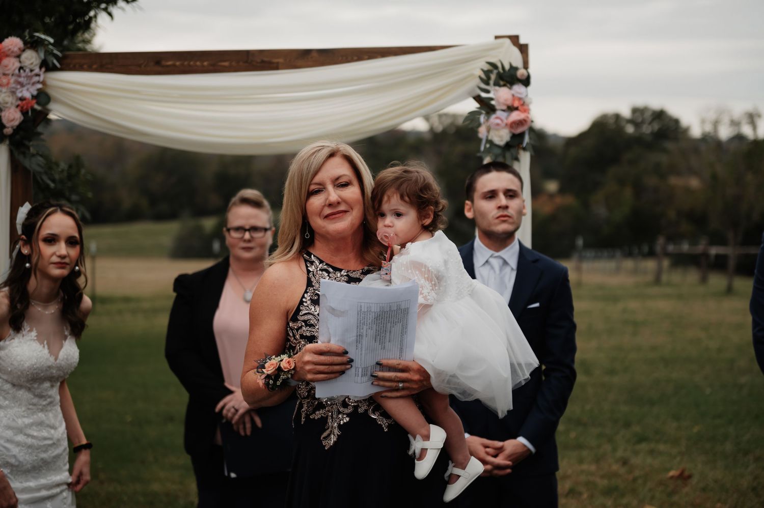 Woman holding toddler in white dress at outdoor wedding ceremony with bridal party and floral arch backdrop.