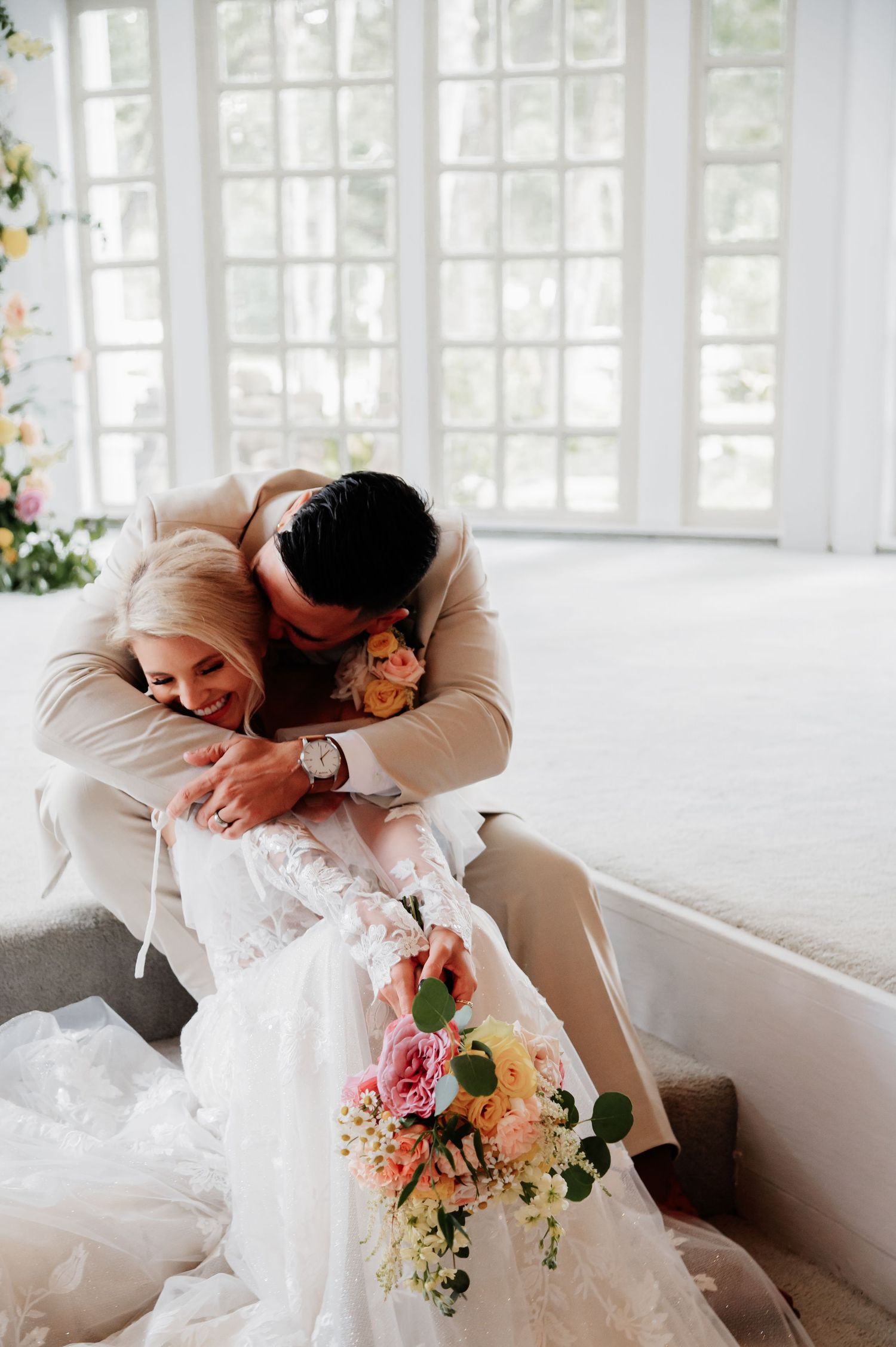 Bride in lace gown holds colorful bouquet while groom in tan suit embraces her tenderly in bright wedding venue.