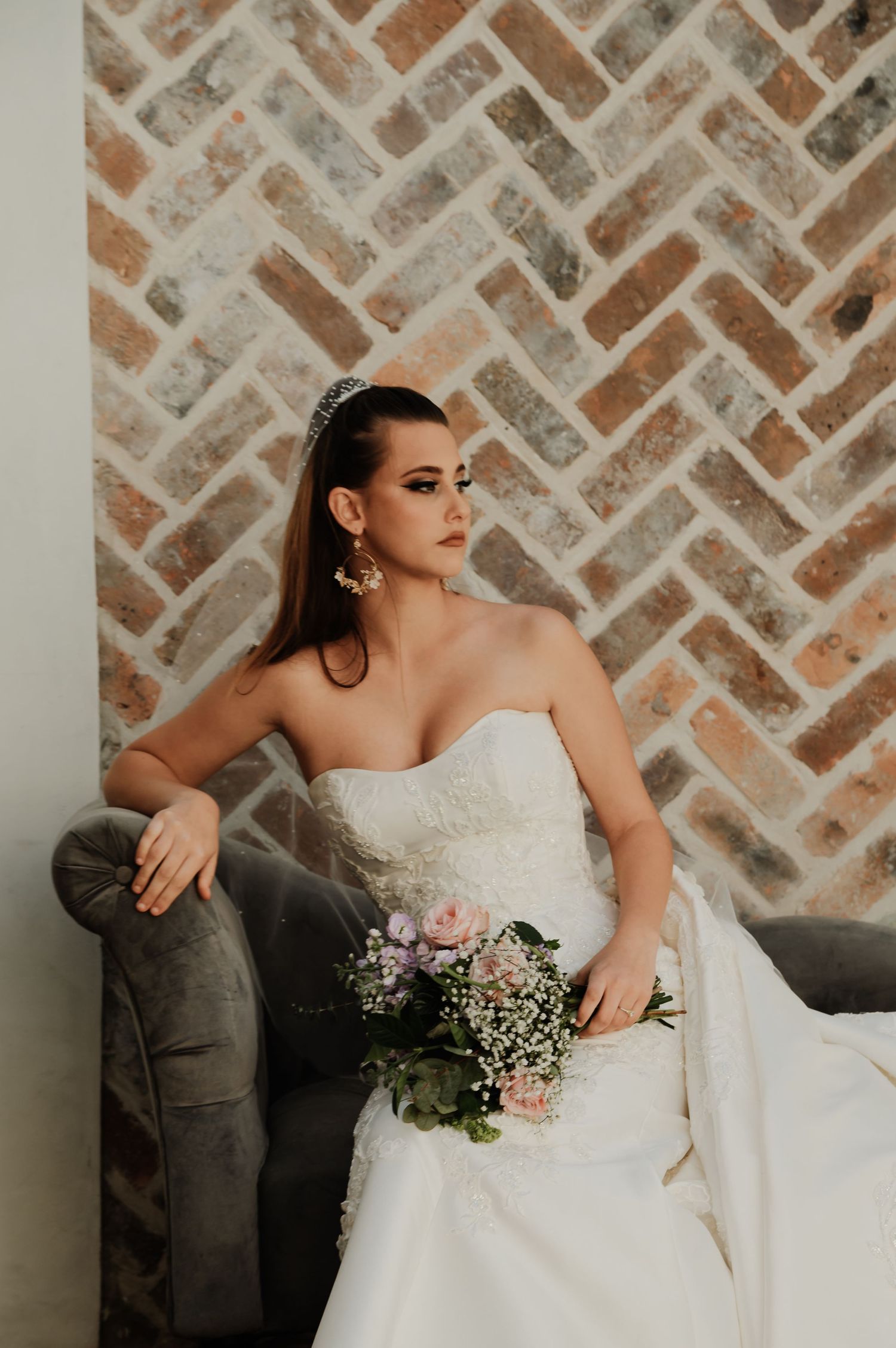 Bride in strapless white gown holding floral bouquet, seated on gray velvet chair against herringbone brick wall.