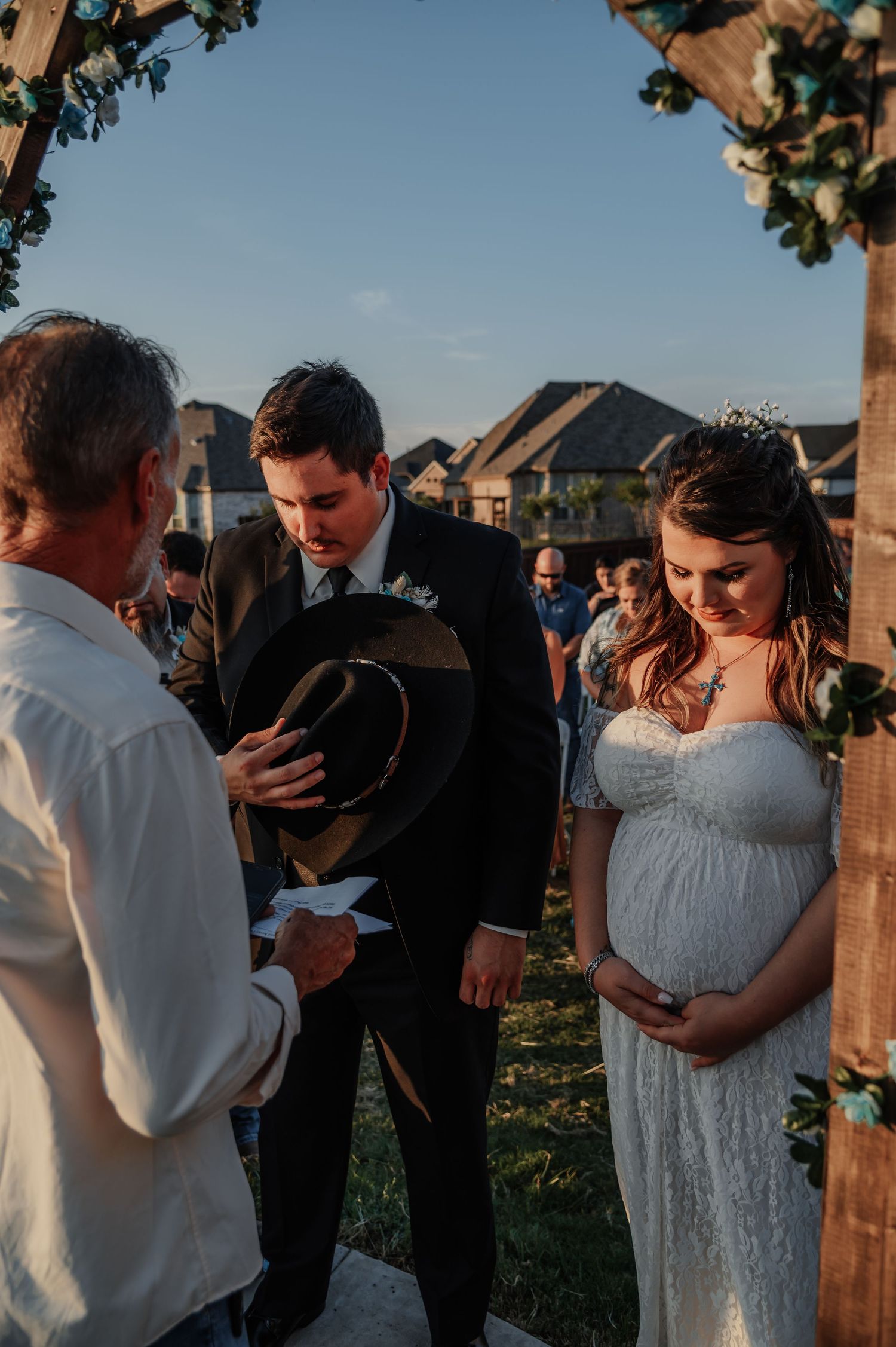 Outdoor wedding ceremony with bride in lace gown and groom holding cowboy hat, bowing heads in prayer under floral arch.
